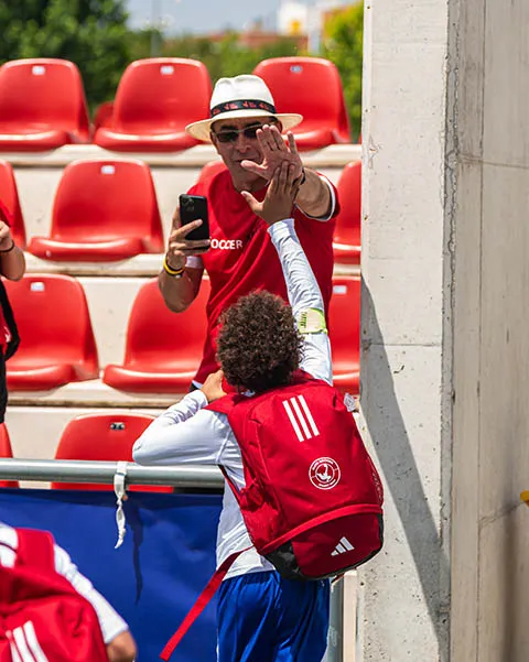 A man wearing a white hat and sunglasses gives a high five to a soccer player with a red backpack in a stadium with red seats.