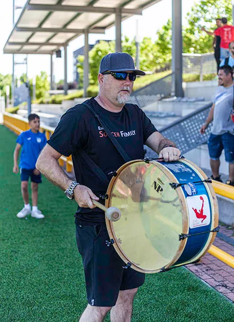Man wearing sunglasses and a cap playing a large drum on a sports field with people in the background.