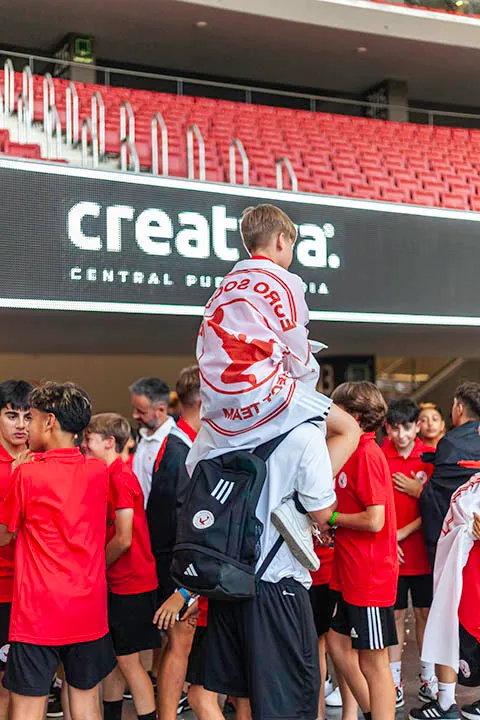 Young boy wearing a white and red flag sitting on an adult's shoulders amidst a crowd of people in red shirts inside a stadium.