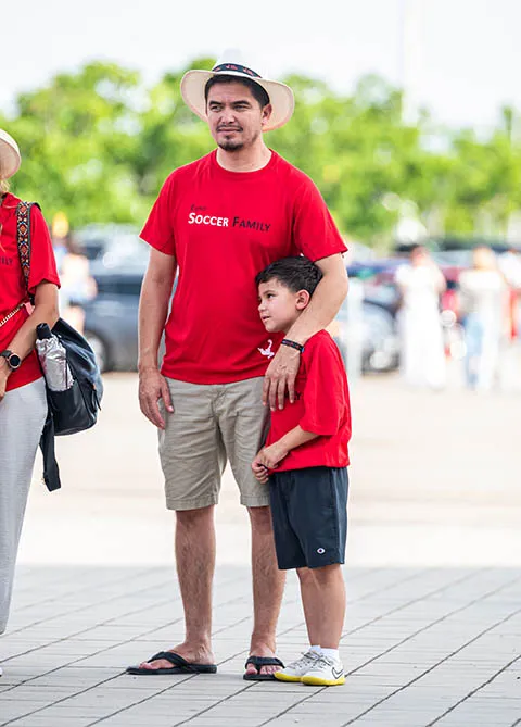 Man wearing a white hat and red 'Soccer Family' shirt standing next to a boy in a matching red shirt outdoors.