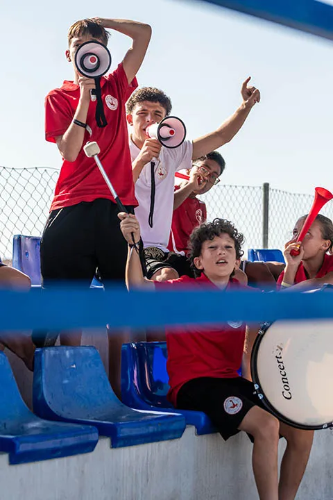 Group of children in red shirts cheering with megaphones, a drum, and a vuvuzela at an outdoor event.