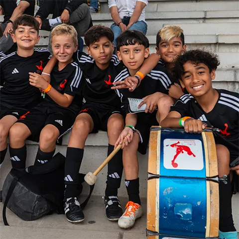 Group of six smiling boys in black sports uniforms sitting on steps, one holding a drum and drumstick.
