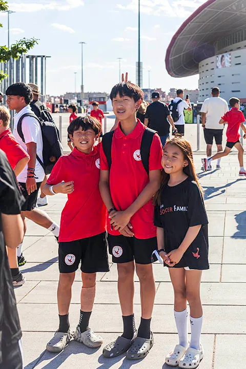 Three children smiling and standing together outdoors; two boys in red soccer uniforms and a girl in a black 'Soccer Family' shirt.