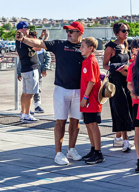 Man in a red cap and black Soccer Family shirt taking a selfie with a boy in a red shirt outdoors on a sunny day.