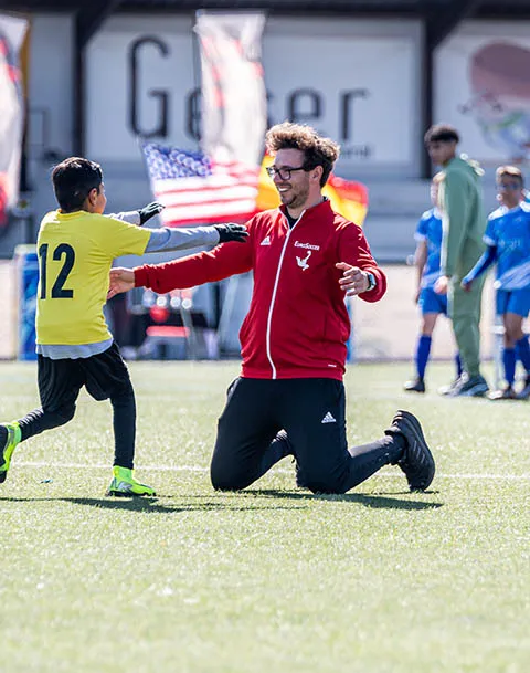 Young soccer player in yellow jersey running to hug a man in a red jacket kneeling on the grass field.