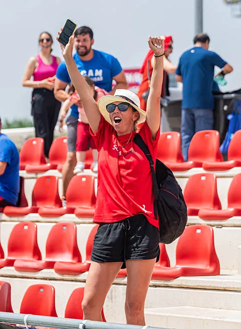 Woman in red shirt and hat cheering with arms raised in stadium seating area.