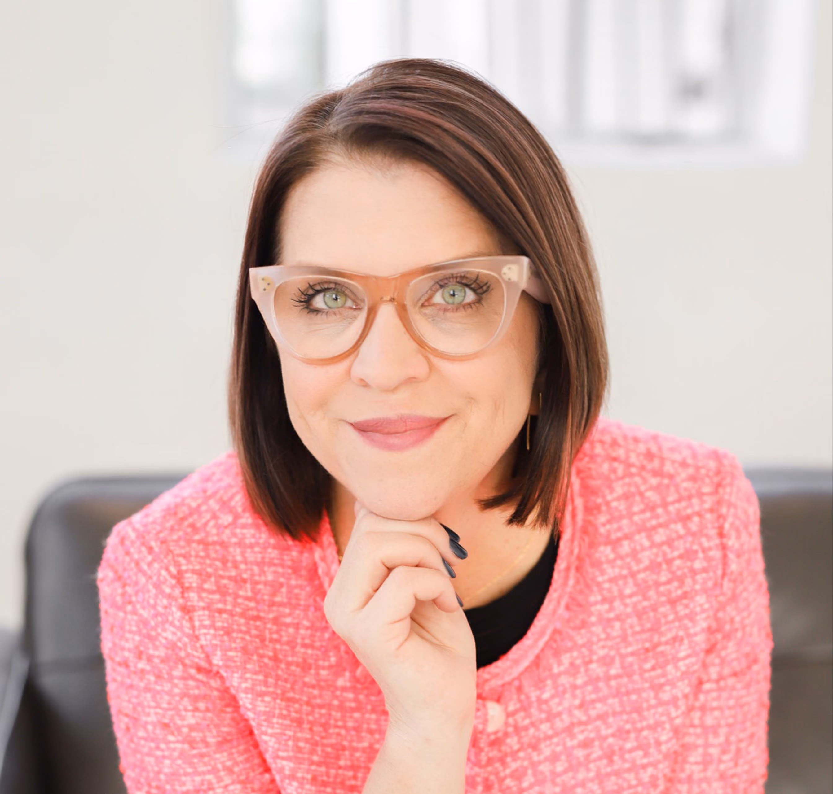 Woman with short brown hair, wearing pink glasses and a pink jacket, resting her chin on her hand.