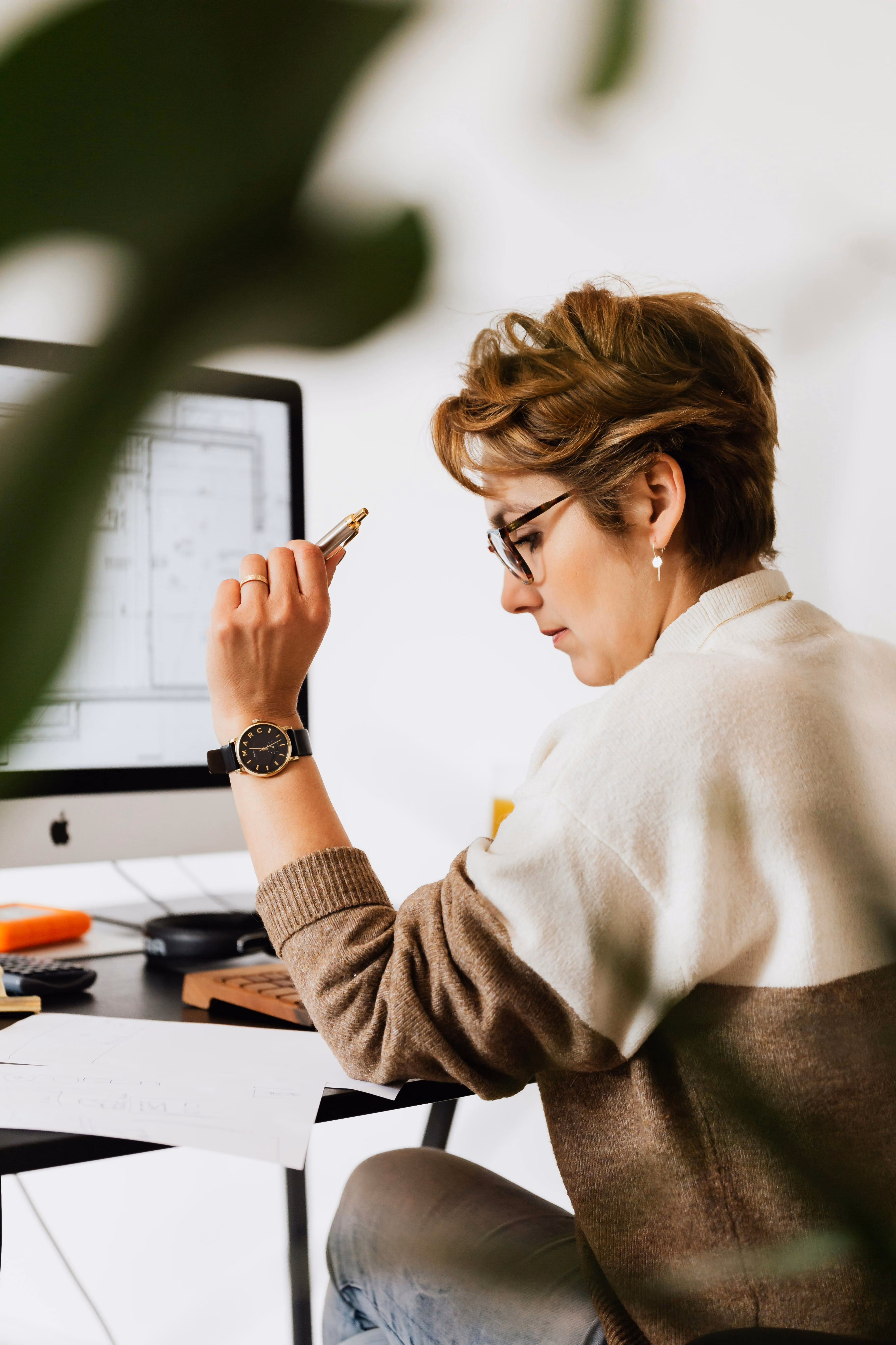 Woman with short hair and glasses working at a desk, holding a pen and looking at a computer screen.