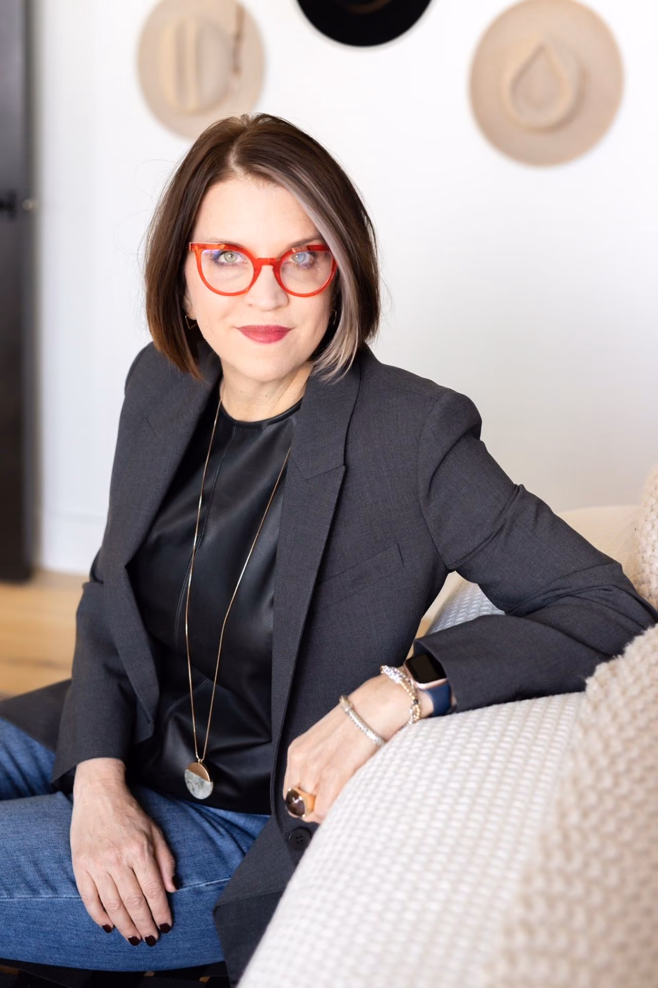 Woman with short brown hair wearing red glasses, gray blazer, and sitting on a beige sofa.
