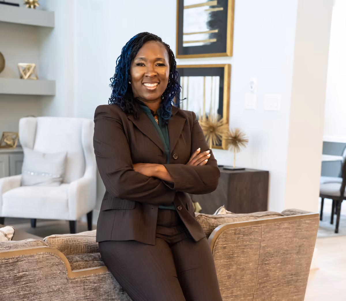 Smiling professional woman in a brown suit sitting on a couch with arms crossed in a modern room.