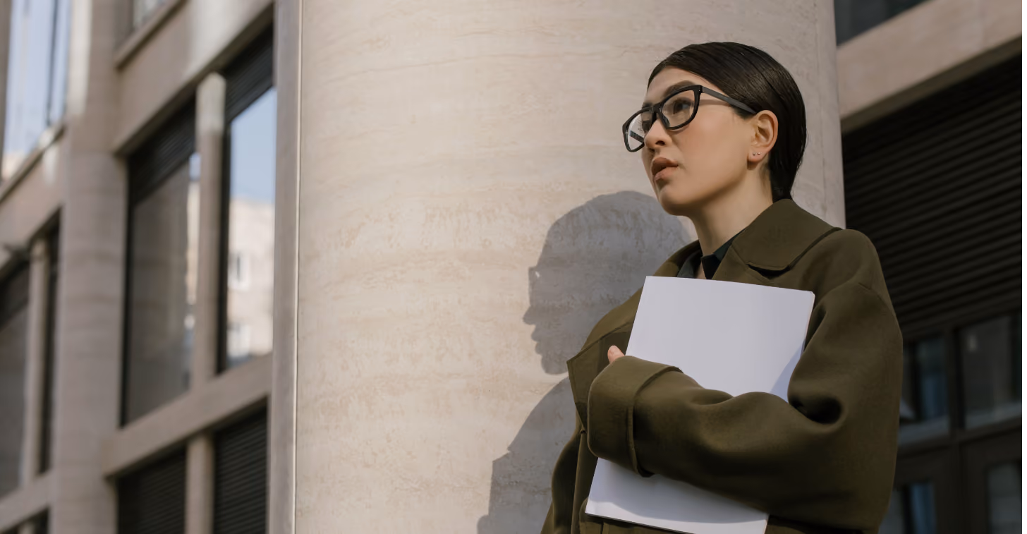 Woman with glasses in green coat holds white folder, leaning against a large stone pillar outdoors.