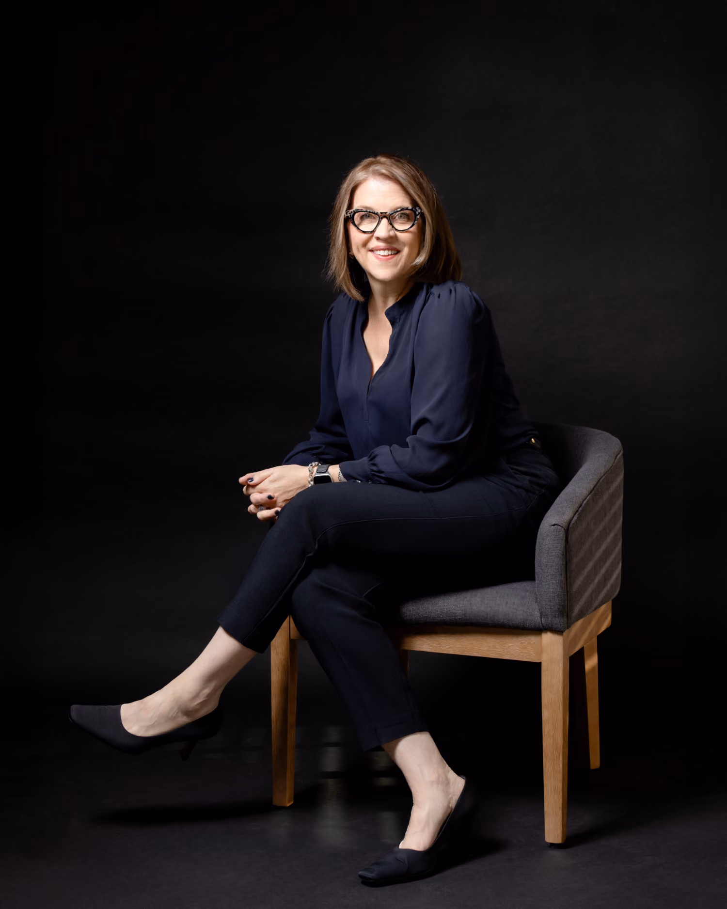 Woman with glasses smiling, seated on a gray chair against a black background.