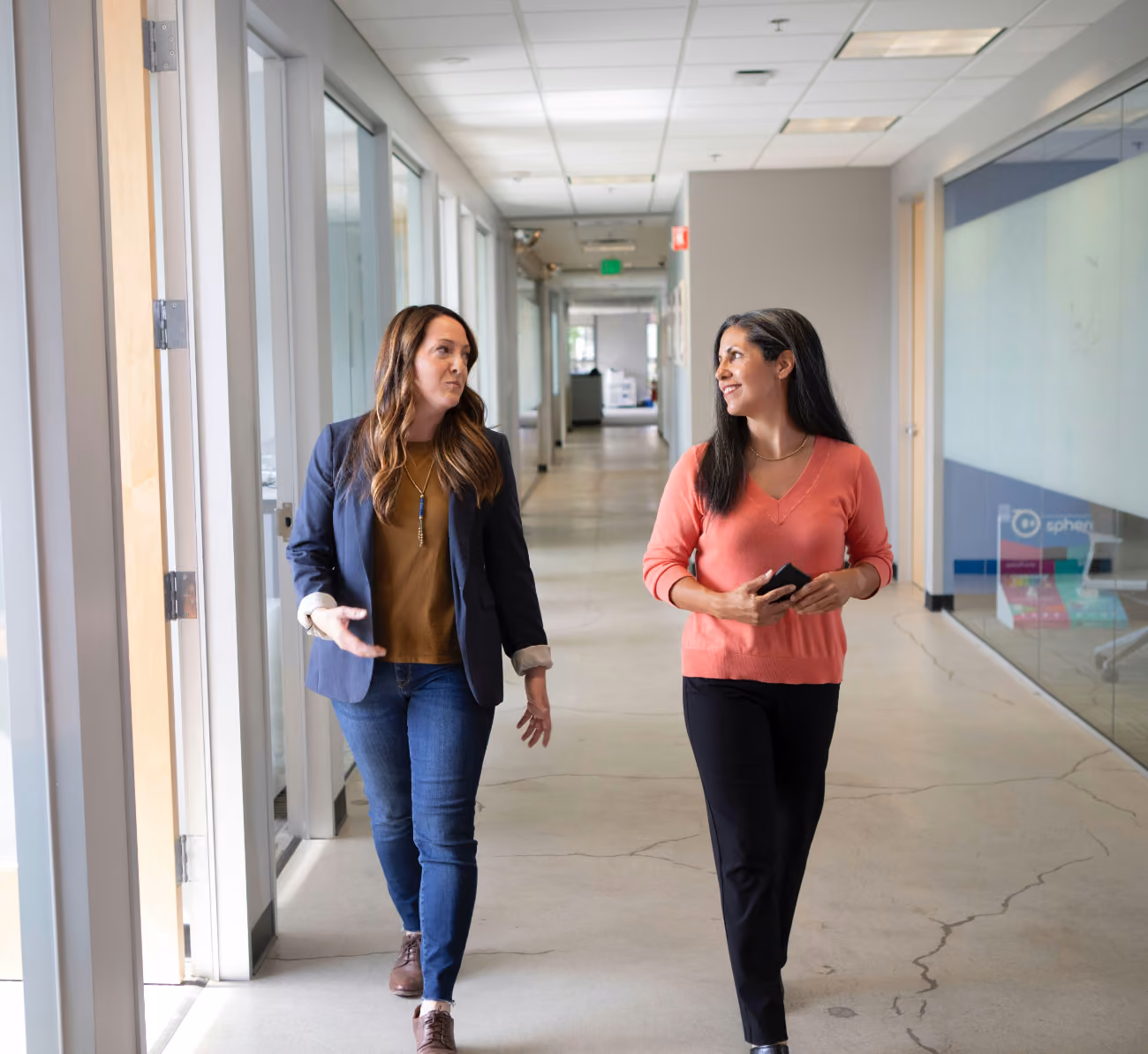 Two women walking and talking in a modern office hallway with glass walls and doors.