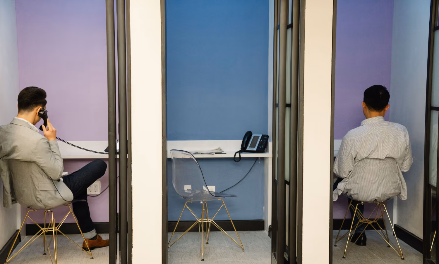 Two men sitting separately in private work booths, one talking on a phone.