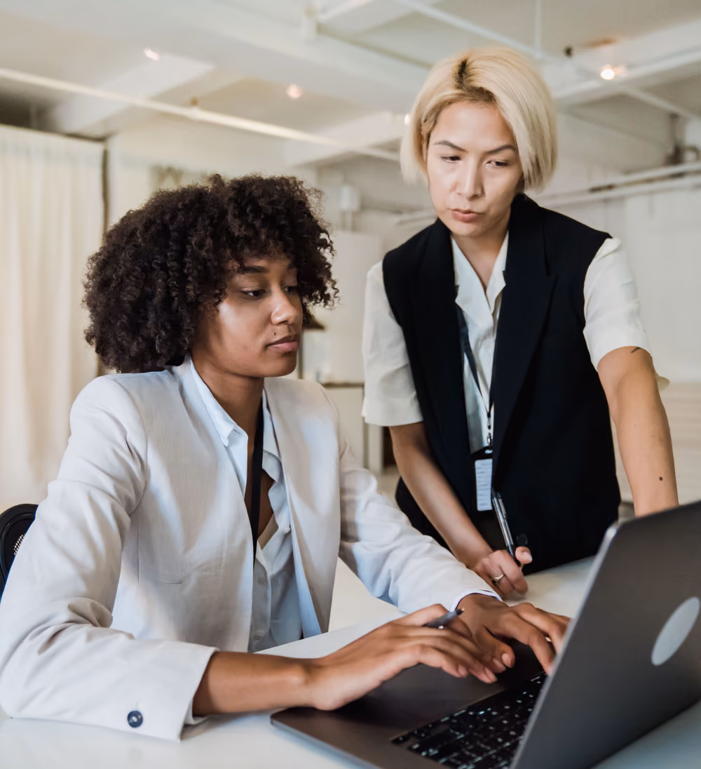Two professional women collaborating and working on a laptop in a modern office setting.