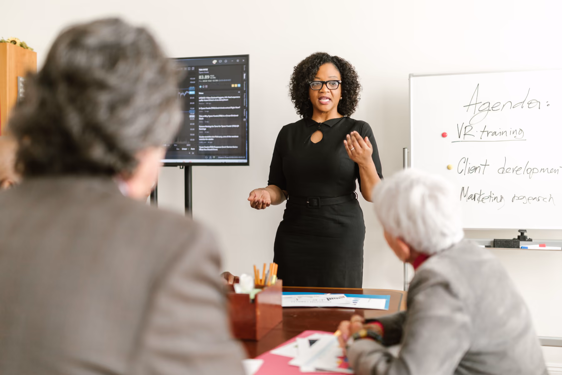 Woman in black dress conducting a meeting with two seated individuals and agenda on whiteboard.