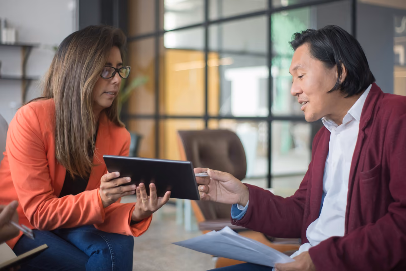 Two business professionals collaborate over a tablet and documents in a modern office setting.