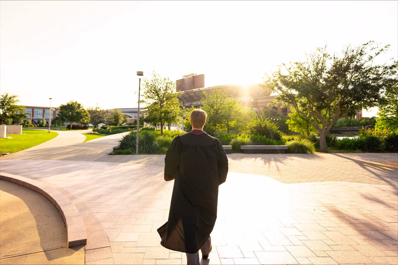 Person wearing graduation gown walking outdoors toward sunlight with trees and buildings in background.