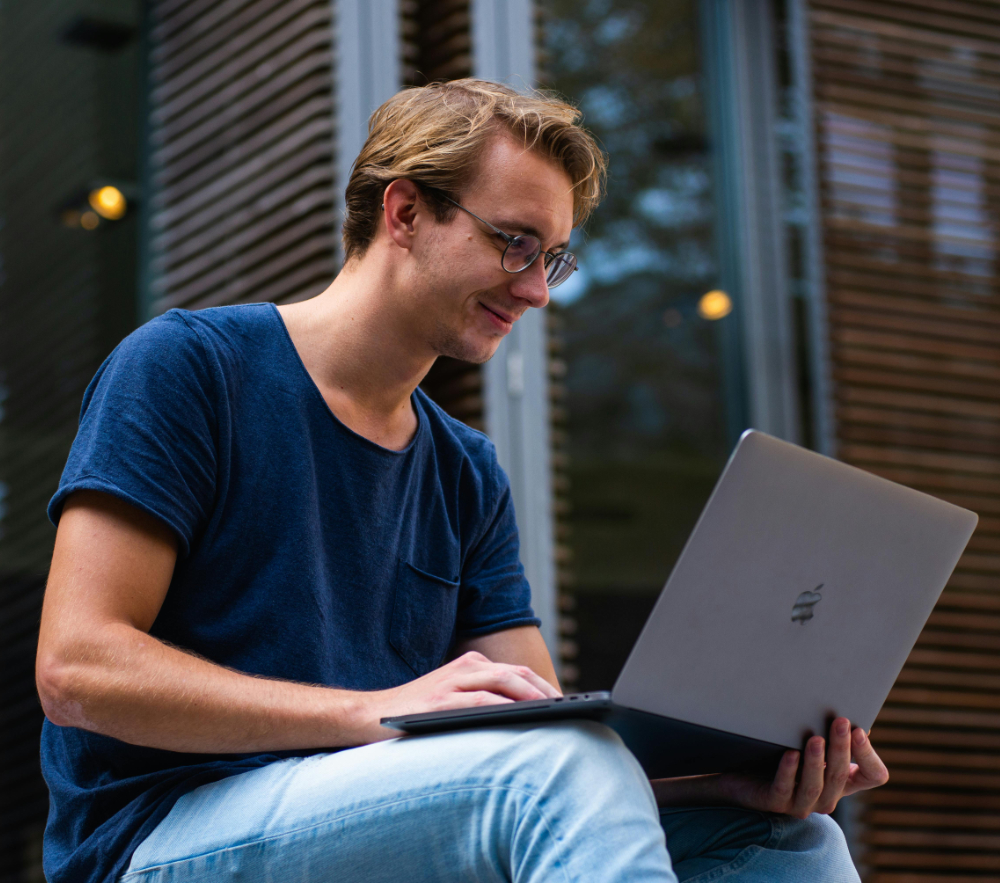Young man with glasses working on a laptop outdoors near a building with window blinds.