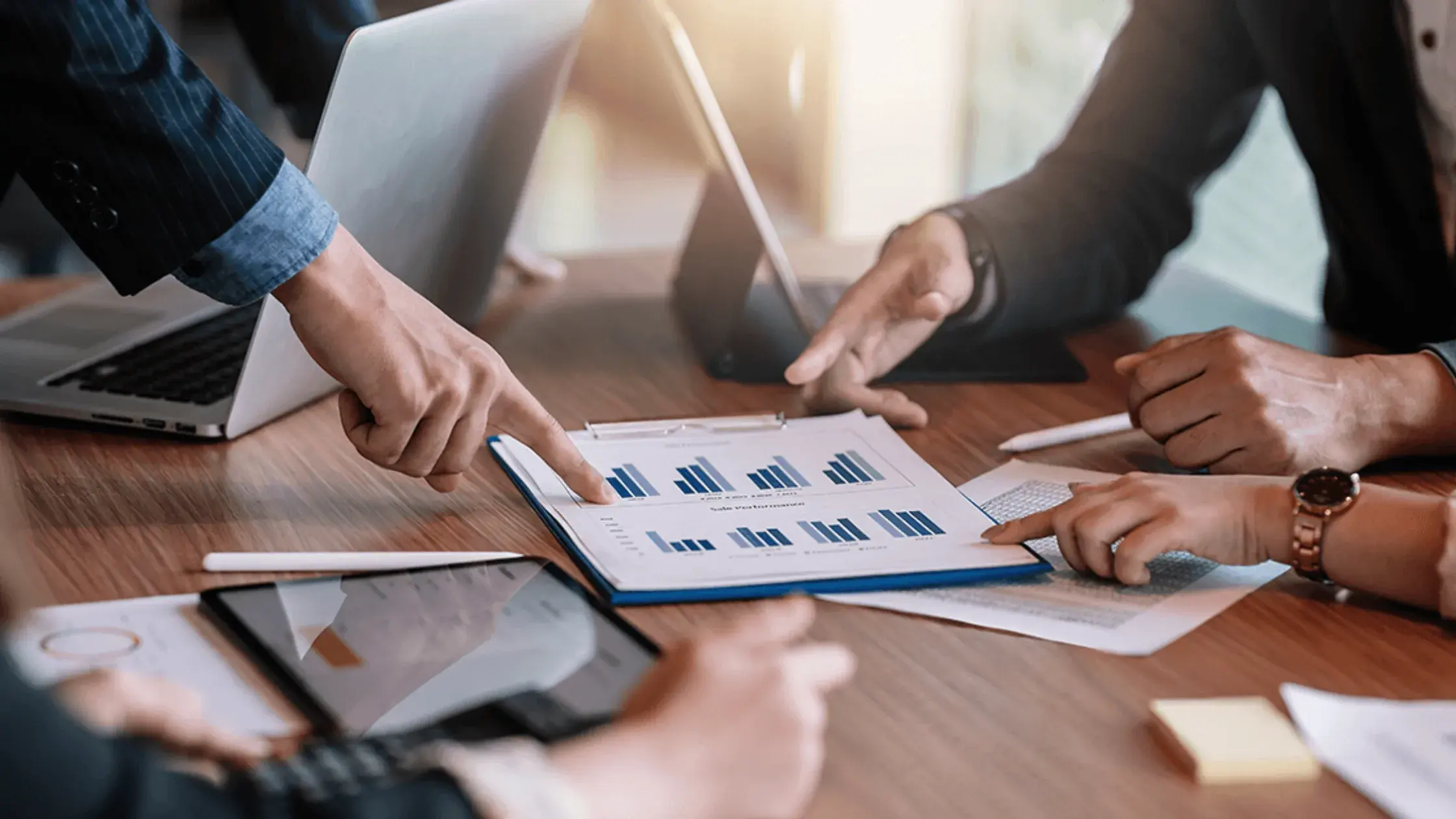 Four people in business attire gathered around a table, reviewing financial charts and using laptops and tablets, suggesting a discussion about business financing options.