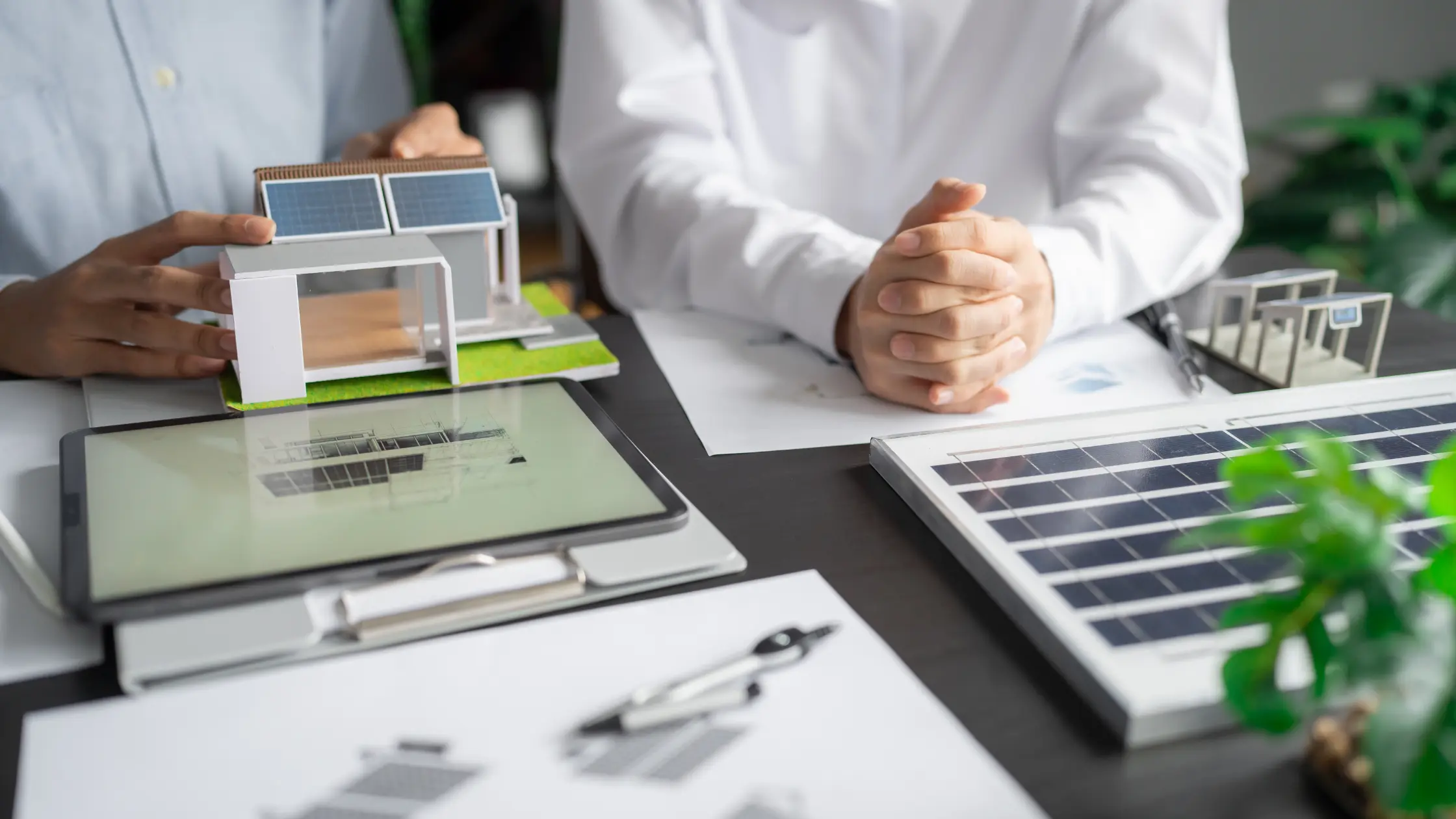  An overhead shot of two people discussing a solar panel project, with a model house and a tablet on the desk highlighting a construction business landscape.
