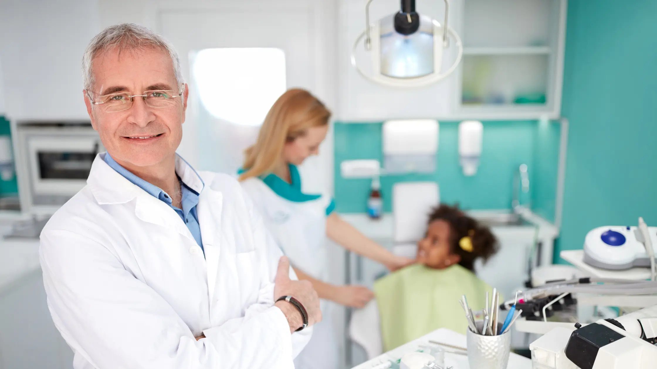A smiling male dentist in a white lab coat stands with his arms crossed, giving a thumbs-up. In the background, a dental hygienist is tending to a young girl in a dentist's chair. Highlighting dental practice loans use.