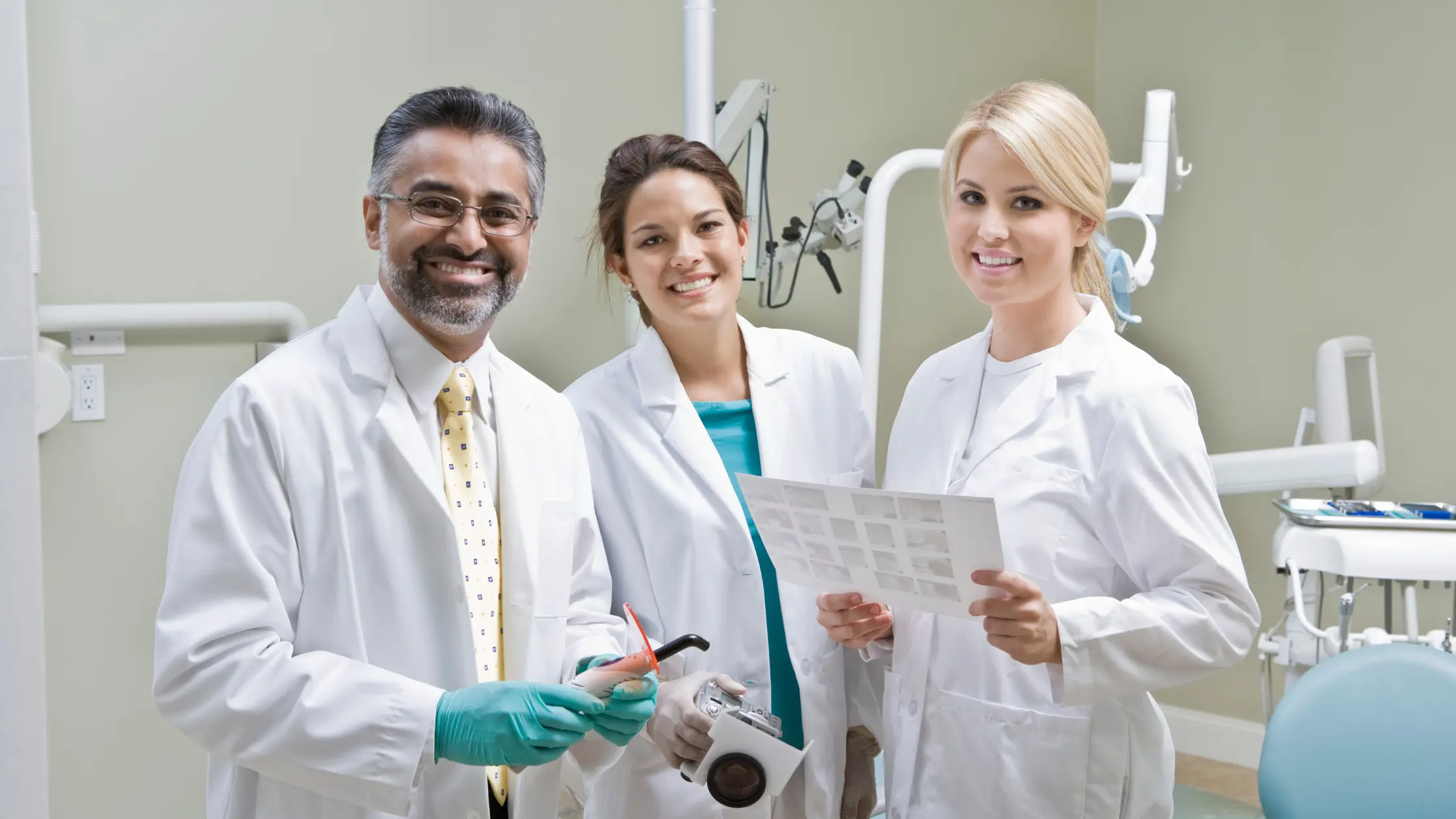  A dentist and two assistants smiling while holding dental equipment and a patient chart.