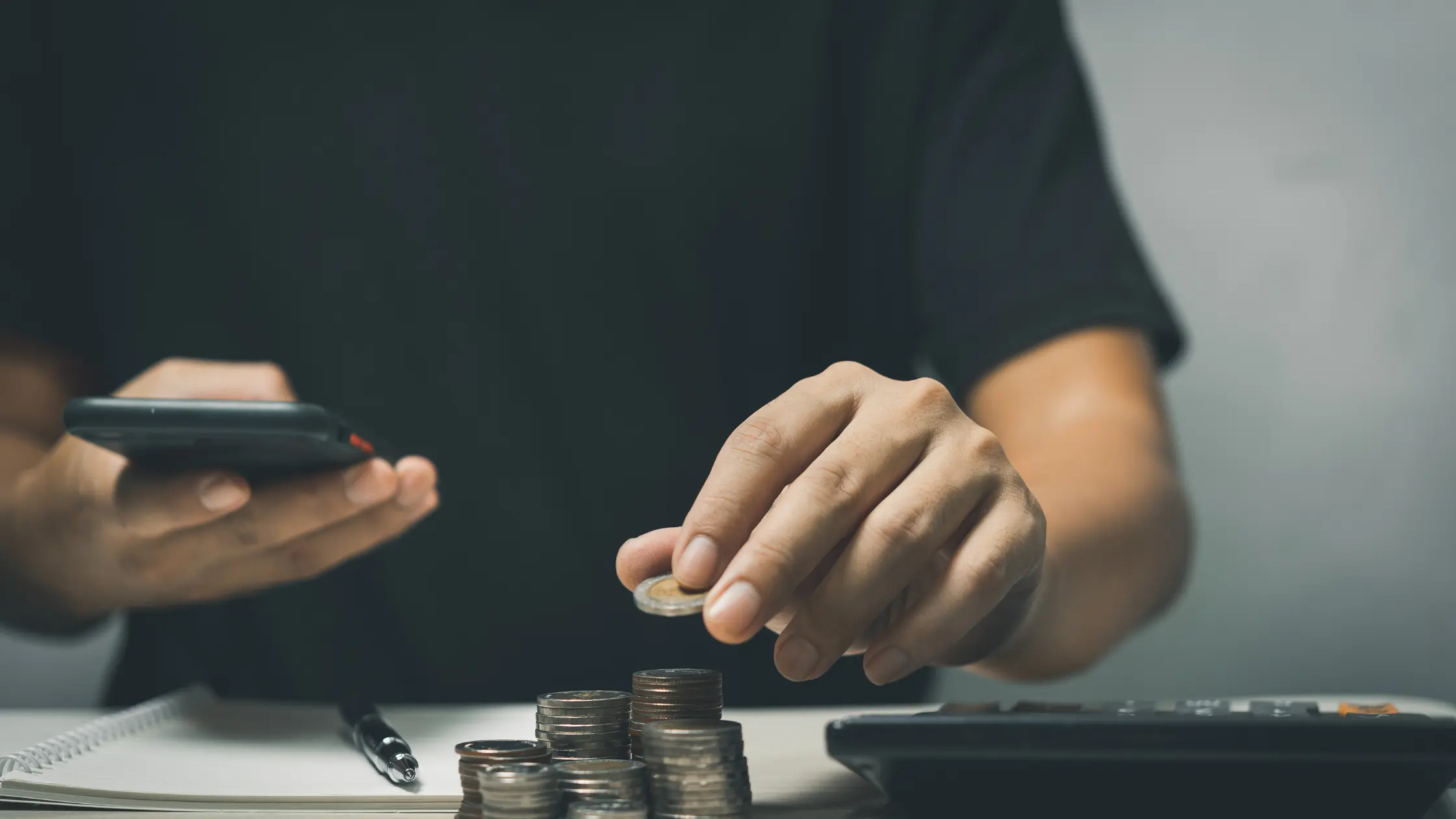 A person holds a smartphone in one hand and places a coin on a stack of coins with the other. A calculator and a notebook are also on the desk. This image suggests themes of factor rates.