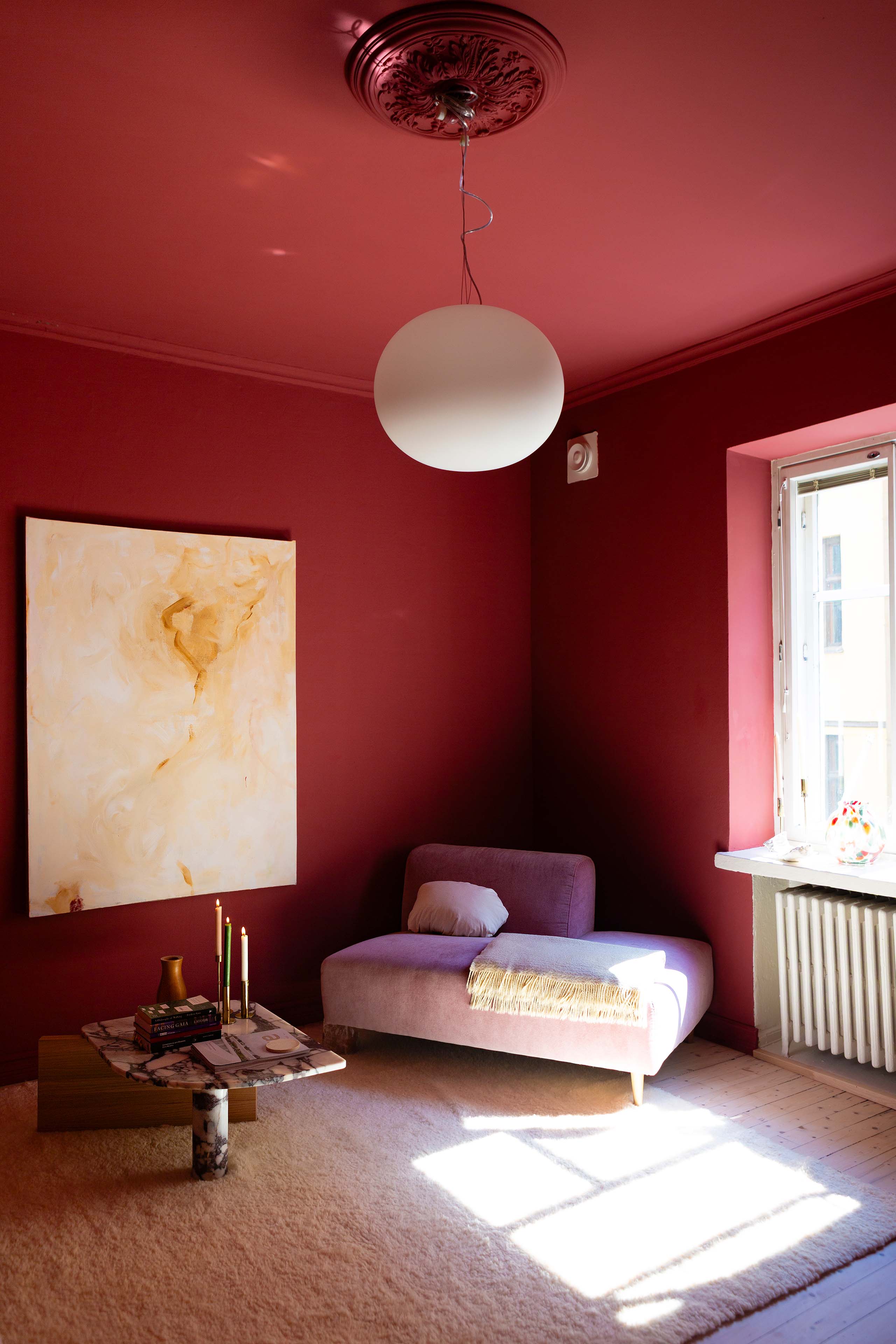 Cozy room with deep red walls, a lavender chaise lounge with a pillow and fringed throw, marble coffee table with books and candles, a large abstract painting, and sunlight streaming through a window.