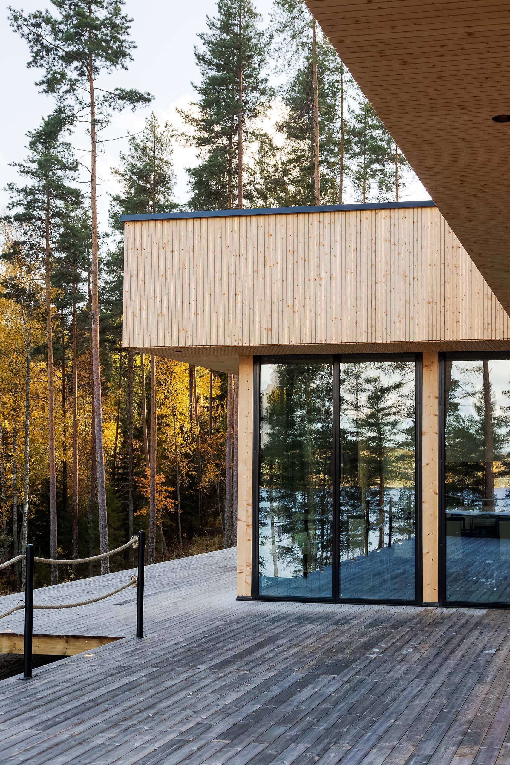 Modern wooden house with large glass windows reflecting forest and lake, surrounded by tall pine trees with autumn foliage.