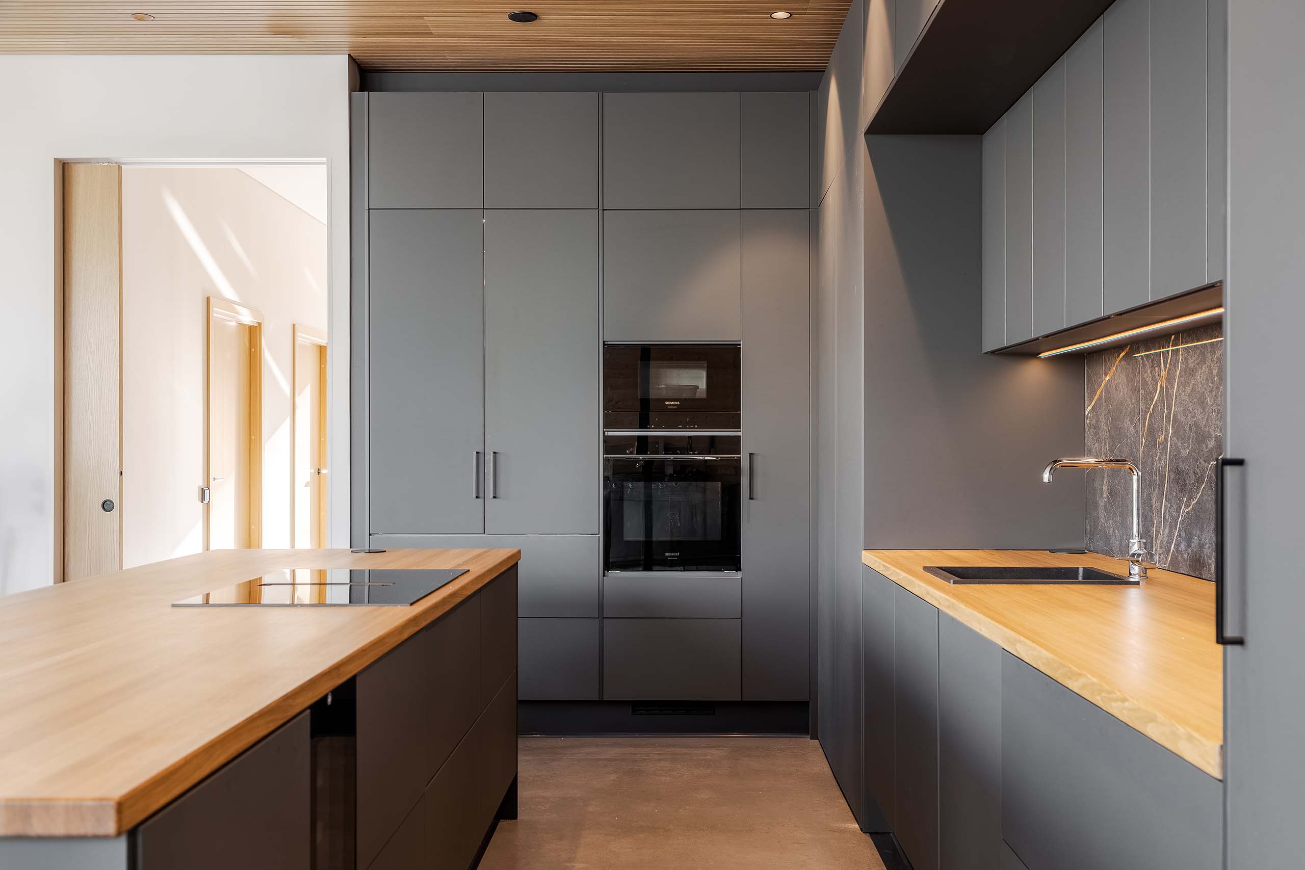 Modern kitchen with matte gray cabinets, wooden countertops, built-in oven, induction cooktop on island, and a stainless faucet with marble backsplash.