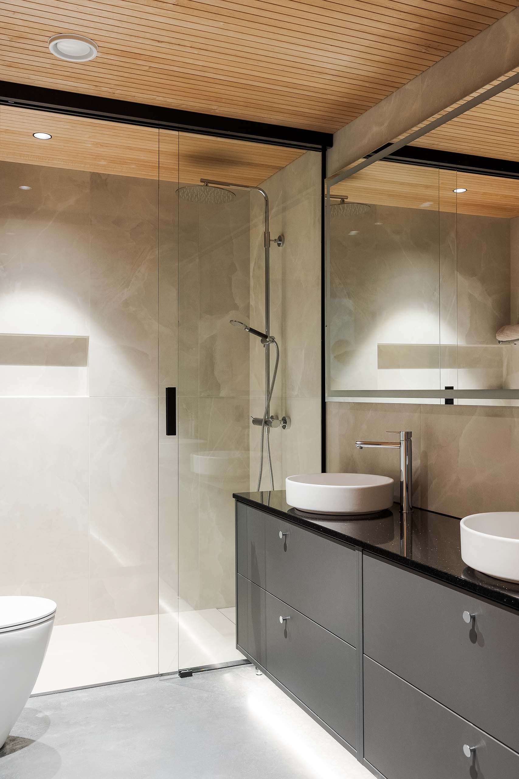 Modern bathroom with glass-enclosed shower, wall-mounted rain showerhead, gray vanity with black countertop, and two round white vessel sinks.