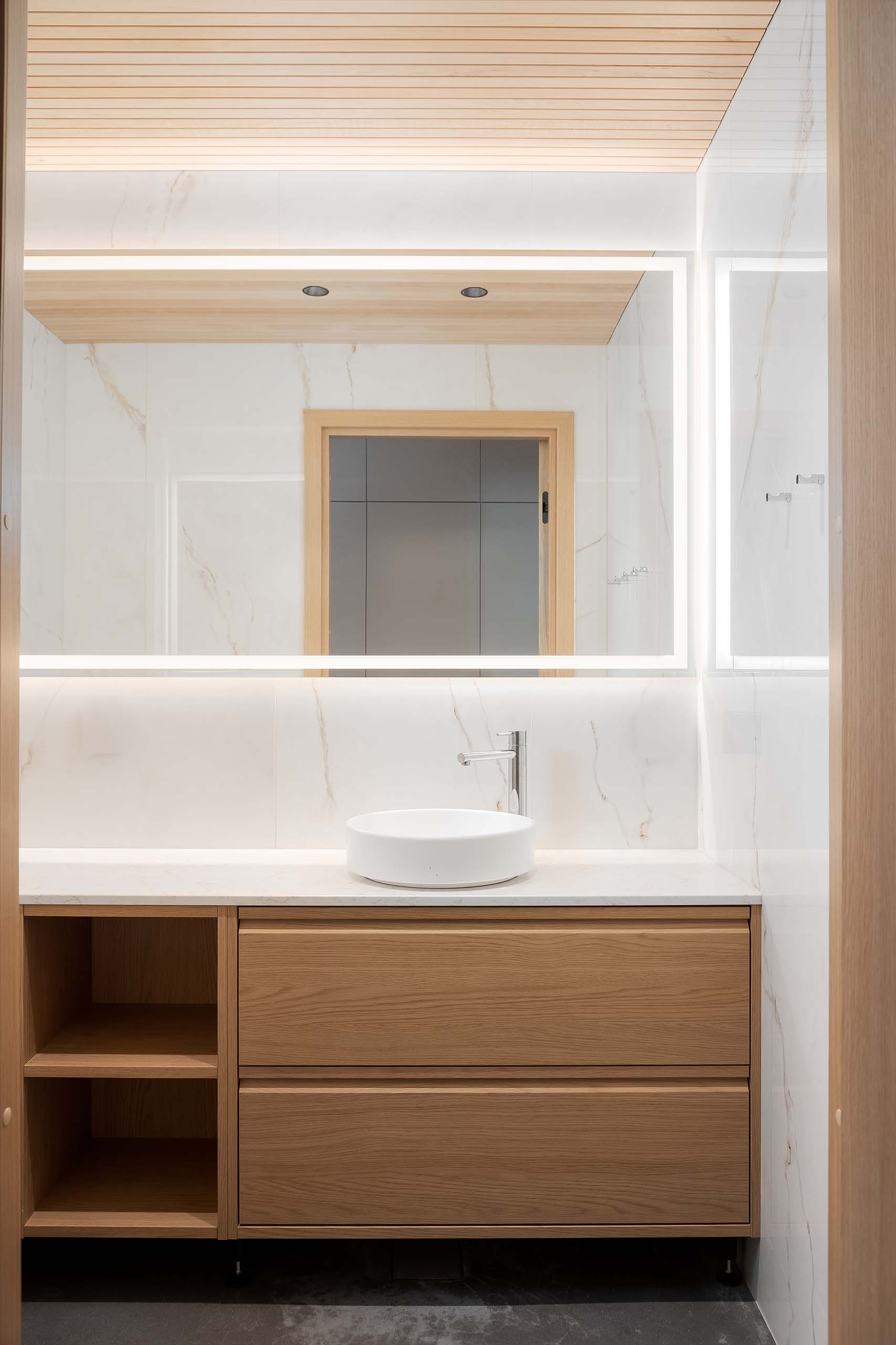 Modern bathroom vanity with wood cabinetry, white round vessel sink, chrome faucet, and large illuminated mirror.