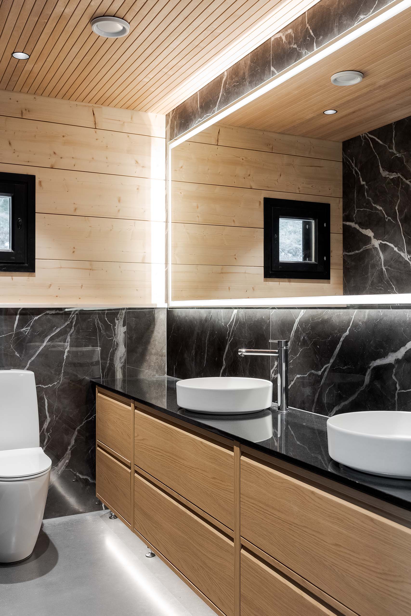 Modern bathroom with wooden cabinetry, black marble backsplash, two white vessel sinks, and a large illuminated mirror.