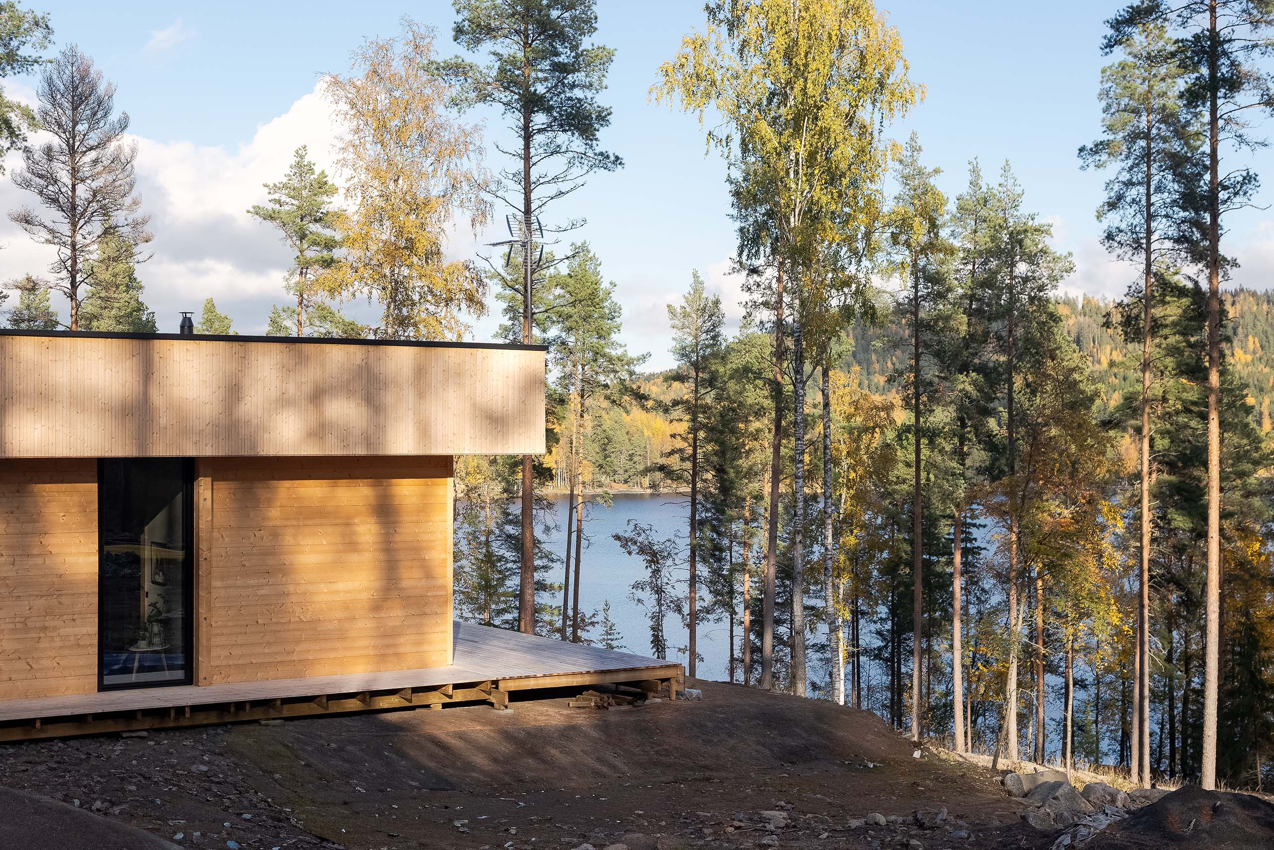 Modern wooden house with large window overlooking a forest and lake in autumn.