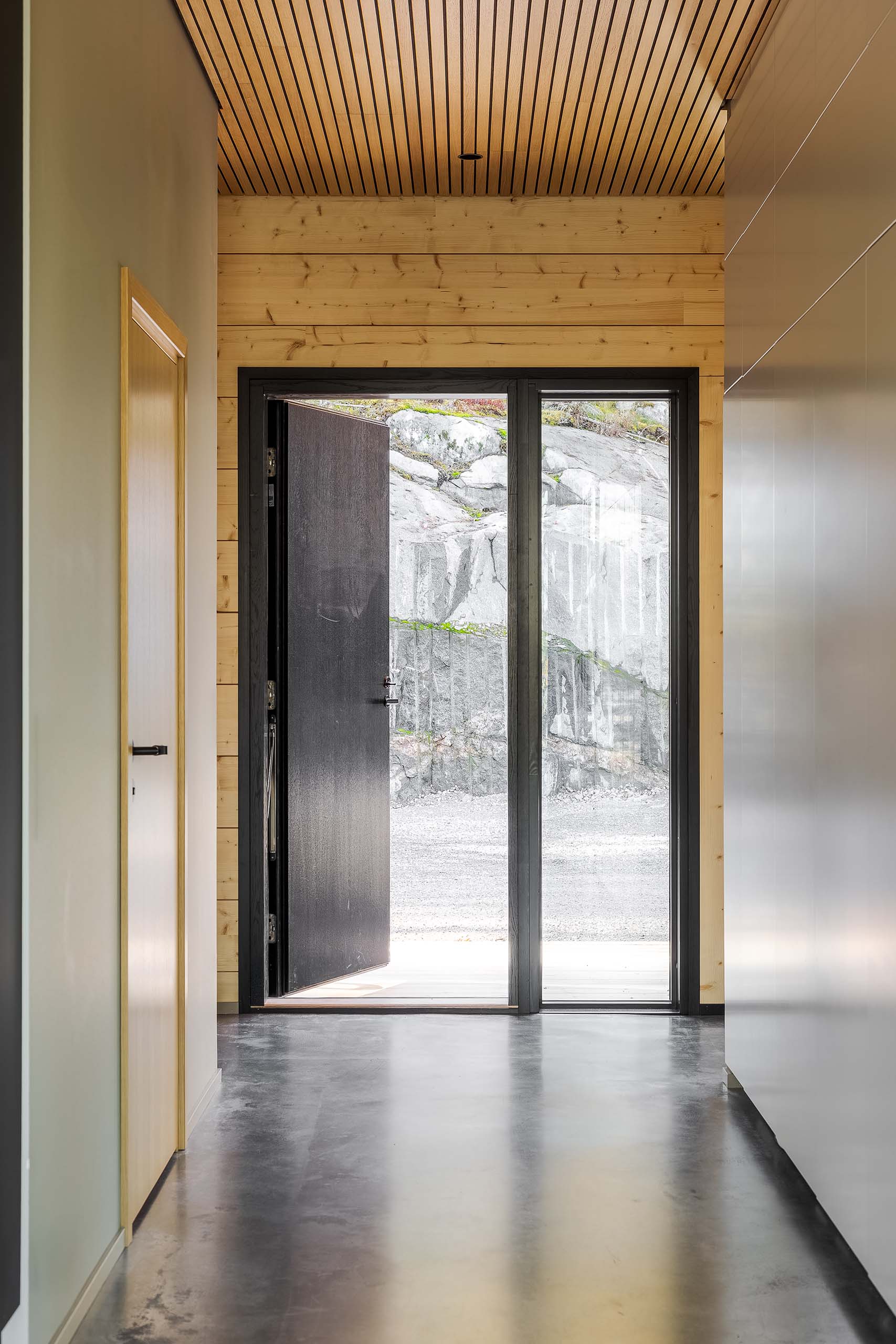 Modern hallway with polished concrete floor, wooden ceiling, and an open black door revealing a rocky outdoor landscape.