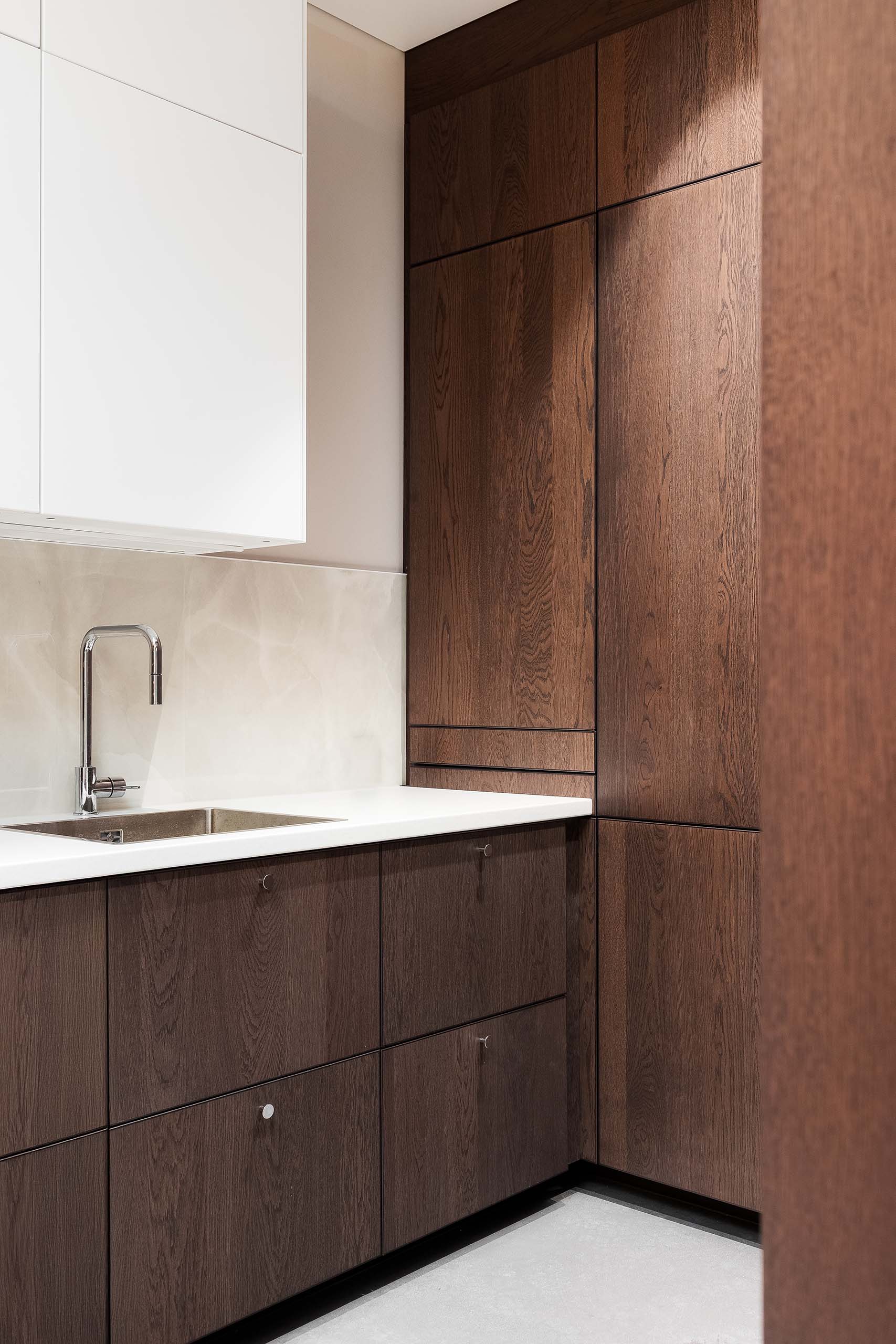 Modern kitchen corner with dark wood lower cabinets, tall wood pantry cabinets, white upper cabinets, white countertop, and a stainless steel sink with chrome faucet.