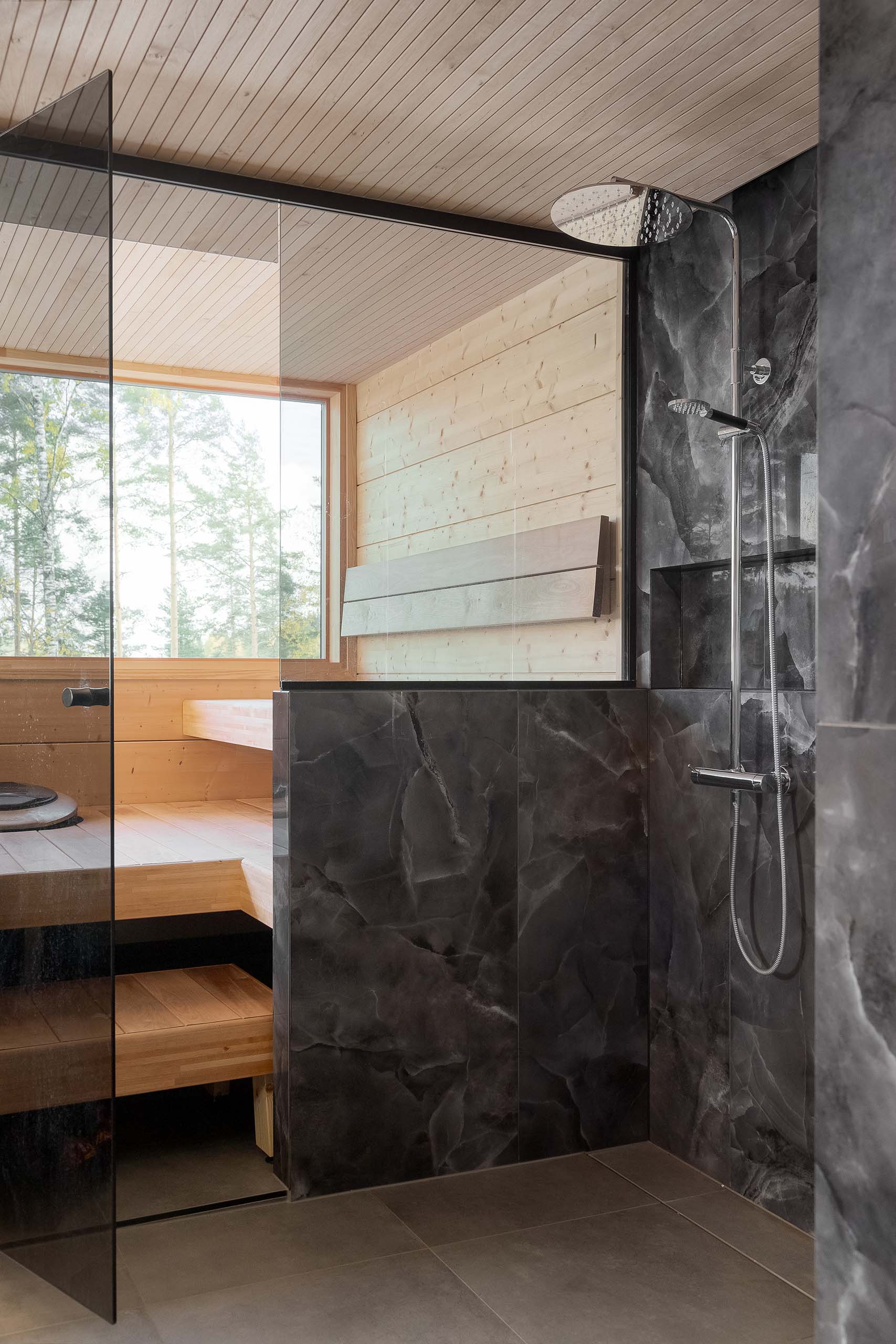 Modern shower area with dark marble tiles and a glass door leading to a wooden sauna with a large window showing trees outside.
