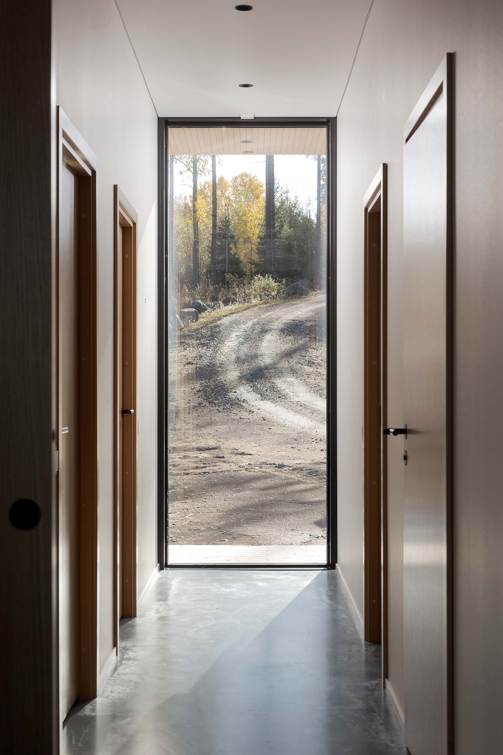 Minimalist indoor hallway with light gray polished concrete floor, light walls, wooden door frames, and a large window showing a dirt road and autumn trees outside.