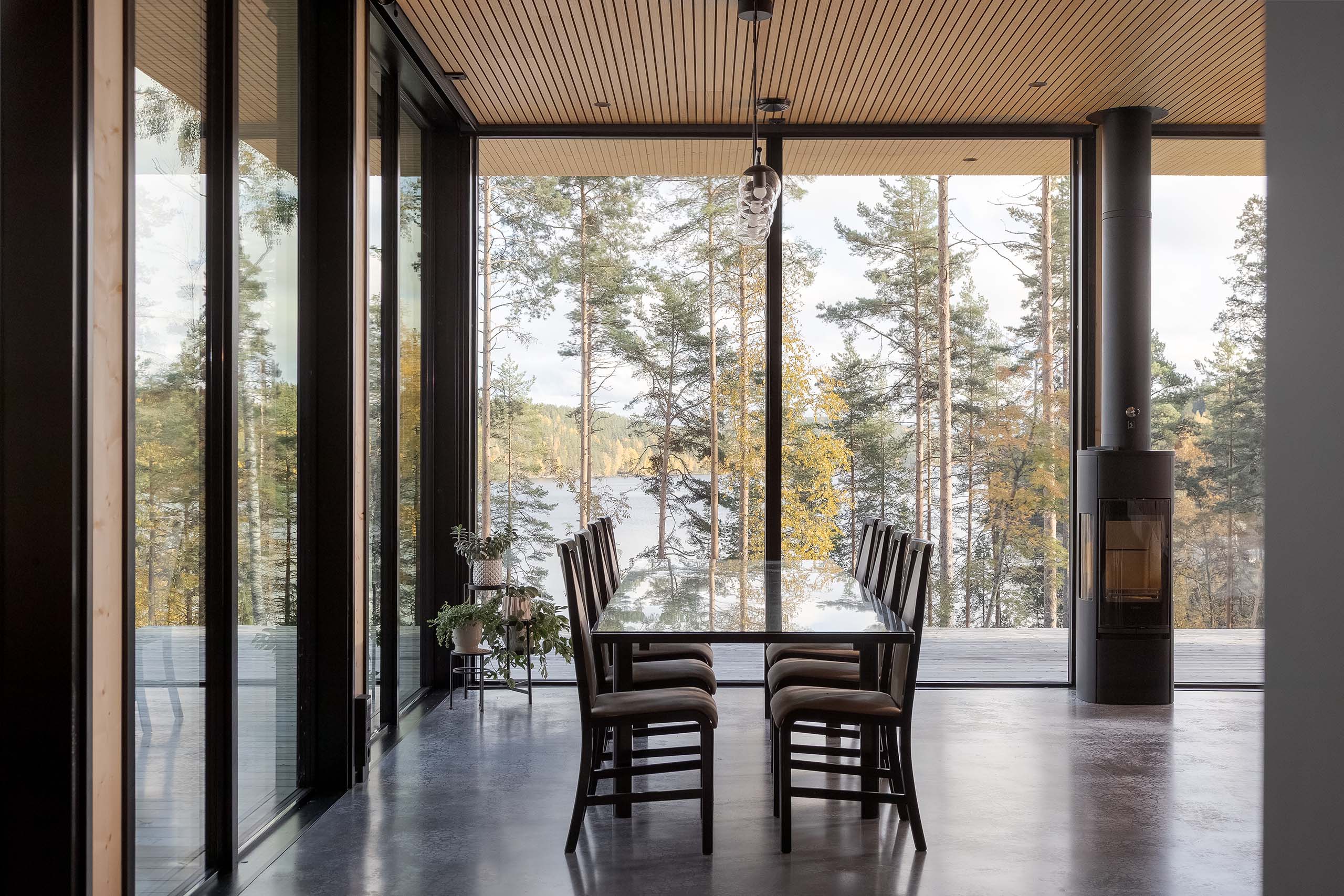 Modern dining area with glass table and eight wooden chairs facing large floor-to-ceiling windows overlooking a forested lake view.
