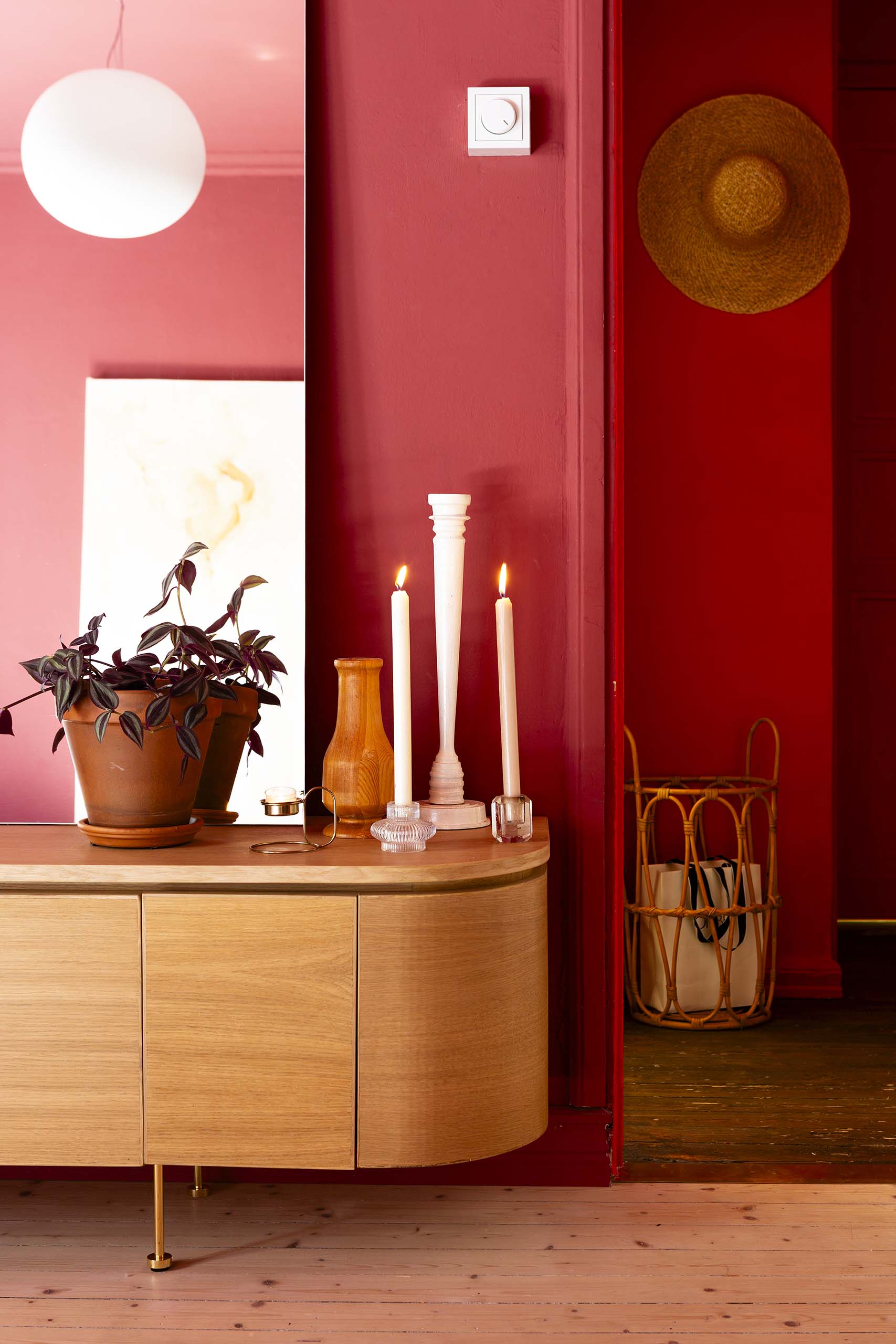 Minimalist wooden sideboard against a red wall, decorated with two lit white candles, a wooden vase, a potted plant, and a large mirror, with a straw hat hanging on the wall near a wicker basket holding bags.