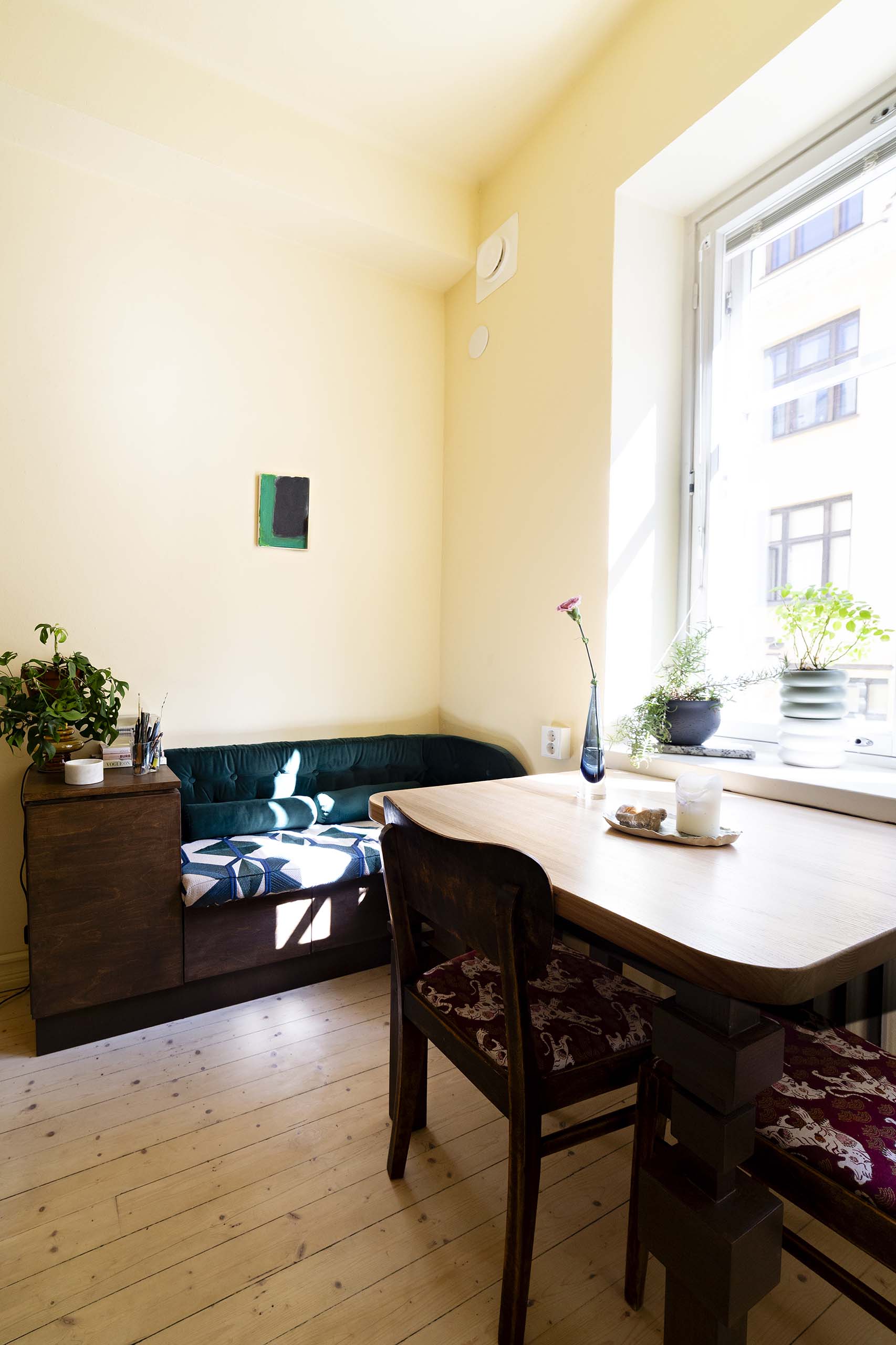 Sunlit cozy corner with a wooden table, two wooden chairs with patterned cushions, a green velvet bench, small plants on a cabinet and windowsill, and a large window showing a building outside.