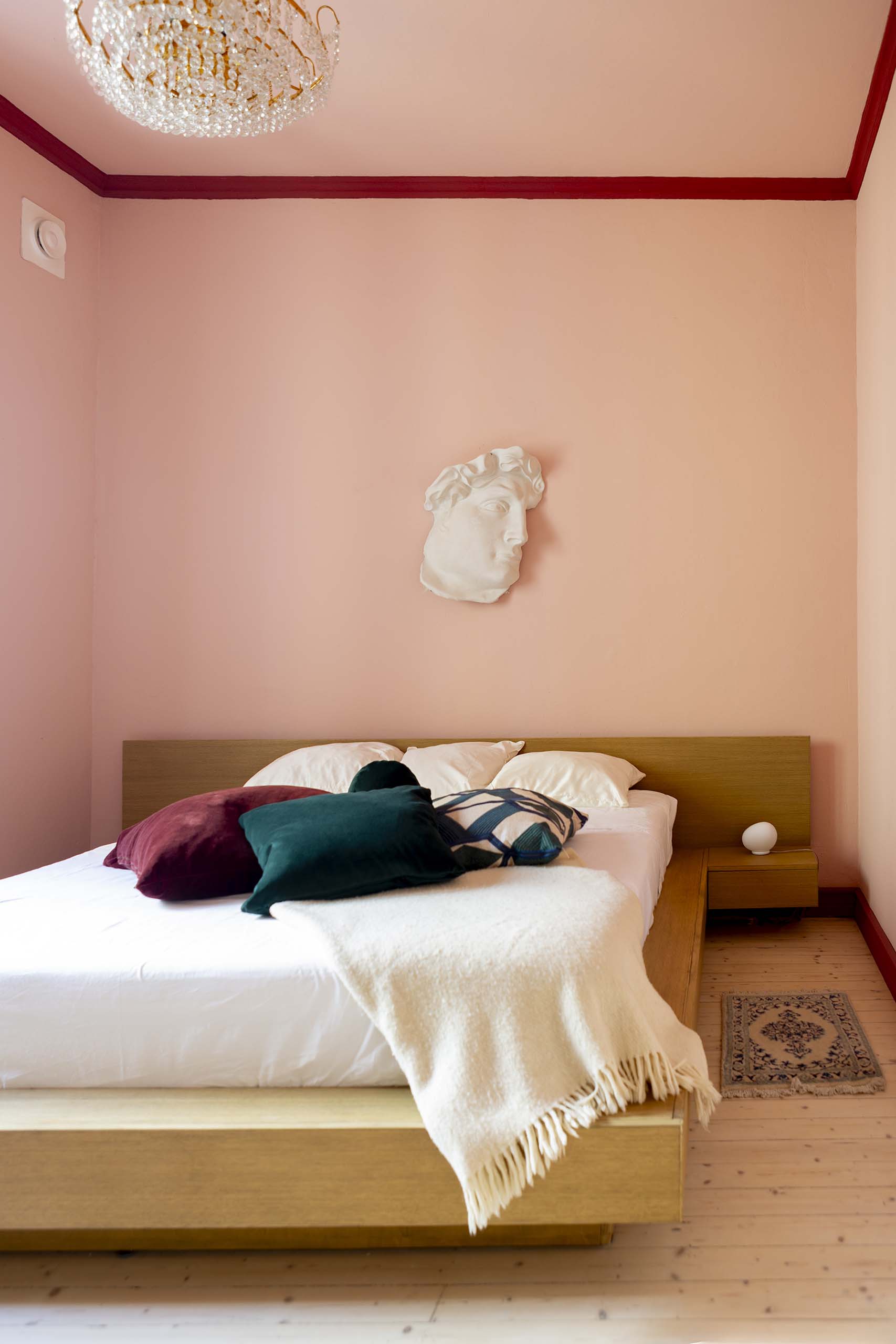 Minimalist bedroom with wooden platform bed, soft pink walls, classical sculpture head on wall, and pillows with white throw blanket.