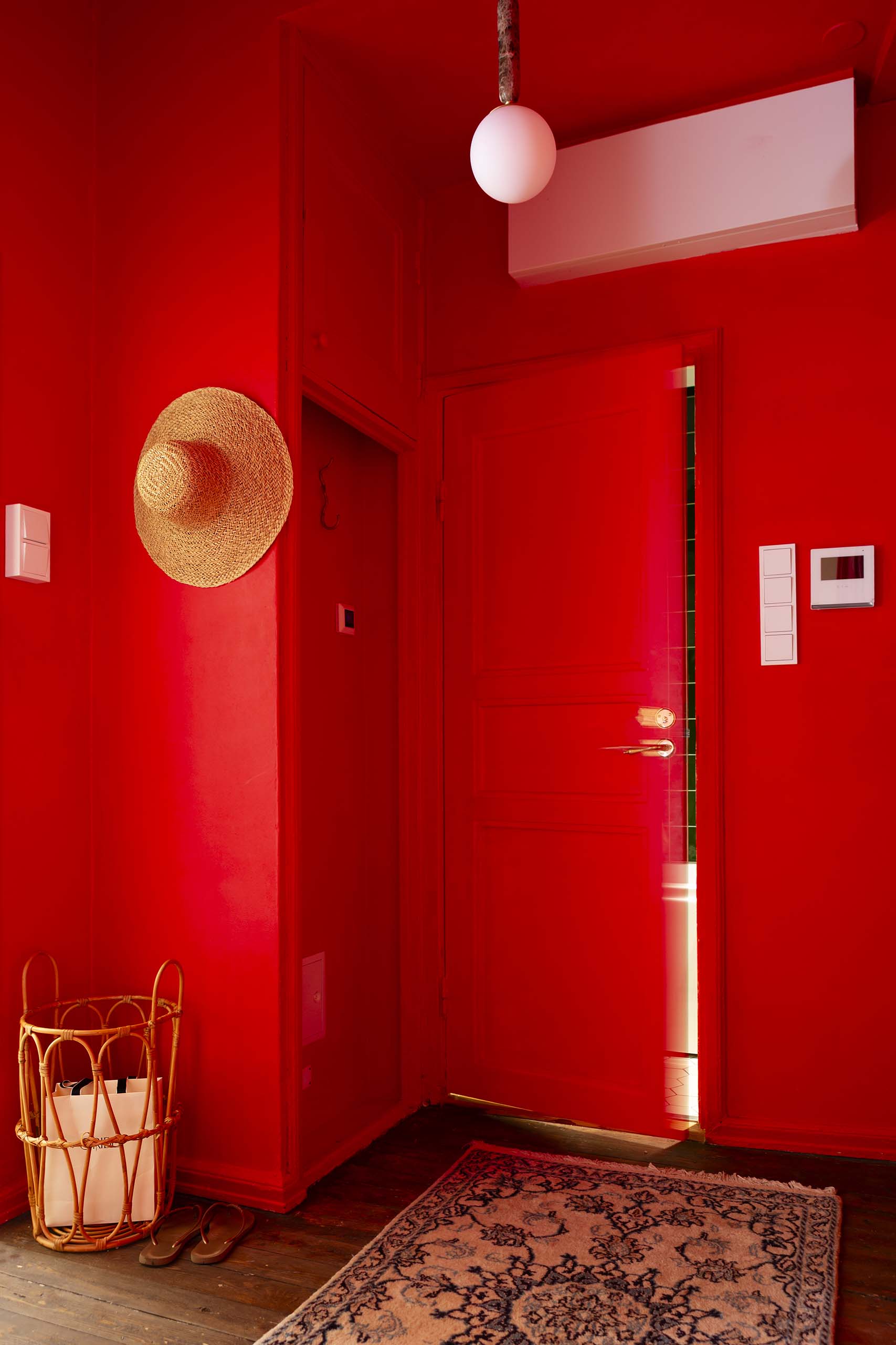 Entrance hallway with bright red walls and door, a straw hat hanging on the wall, a woven basket with bags, flip-flops on the wooden floor, and a patterned rug.