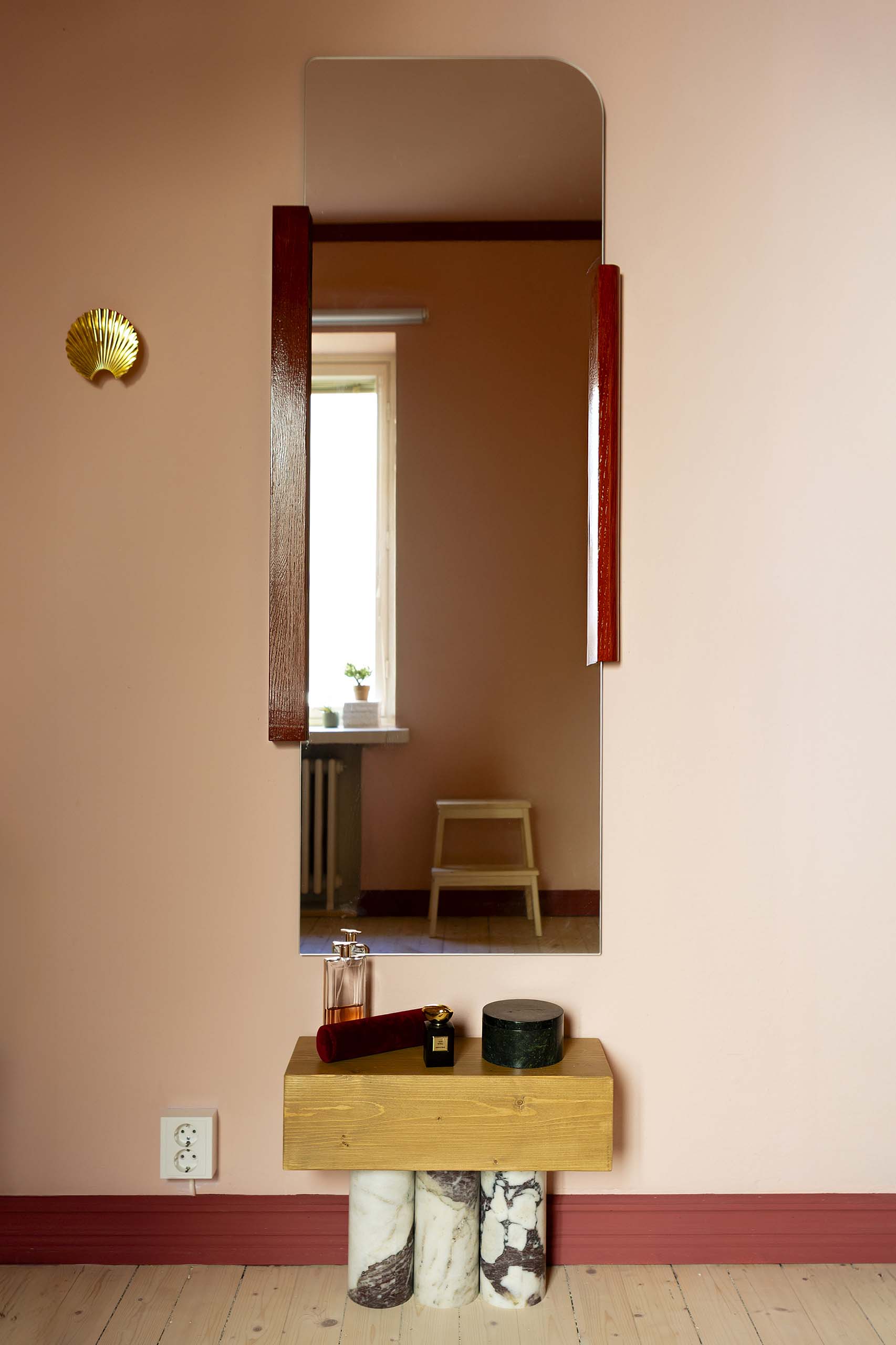 Minimalist vanity setup with a tall rectangular mirror featuring wooden side accents, reflecting a window and stool, and a small table with marble legs holding perfume bottles and a round container.