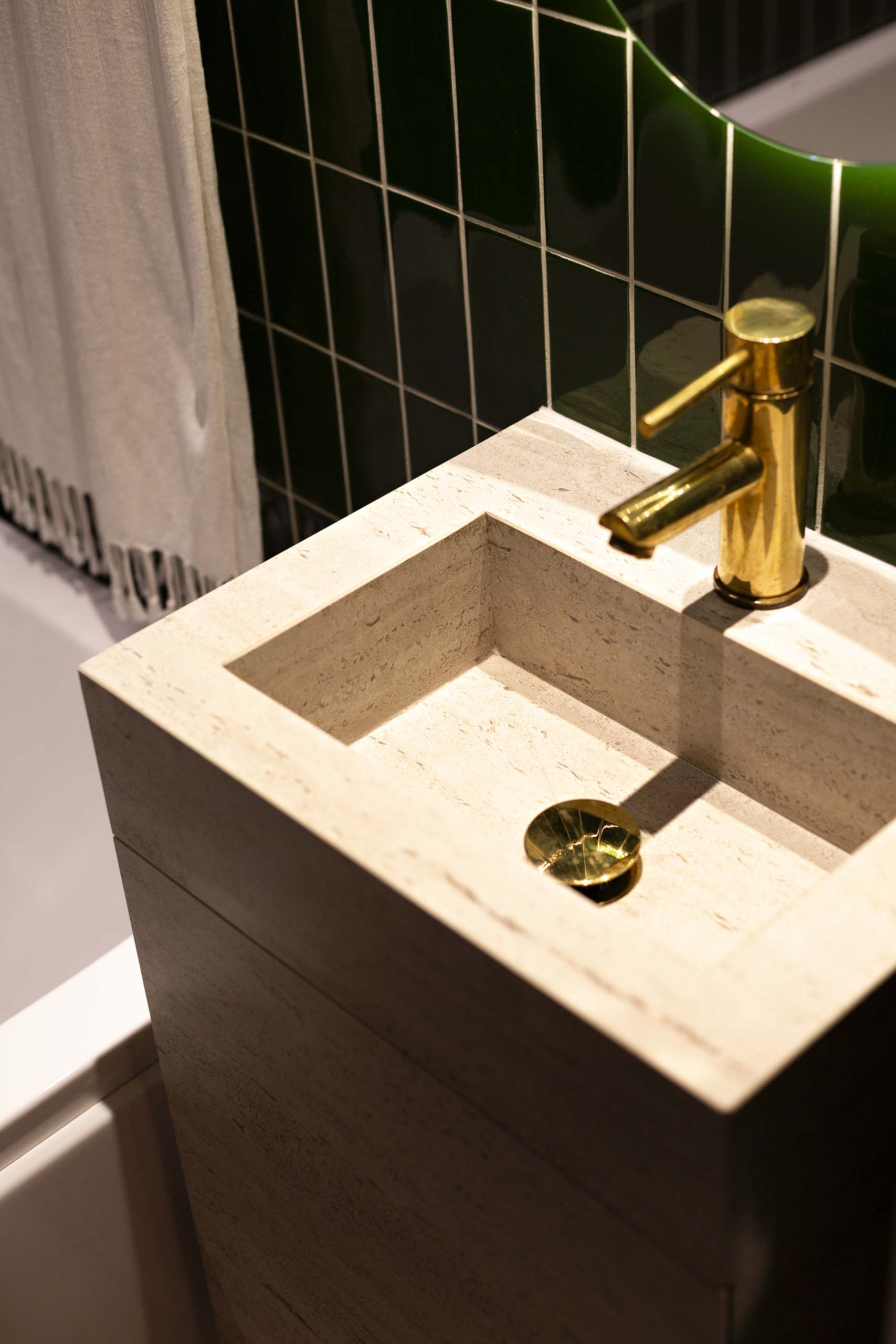 Modern beige stone sink with a gold faucet and drain against dark green tiled wall and a white fringe towel nearby.