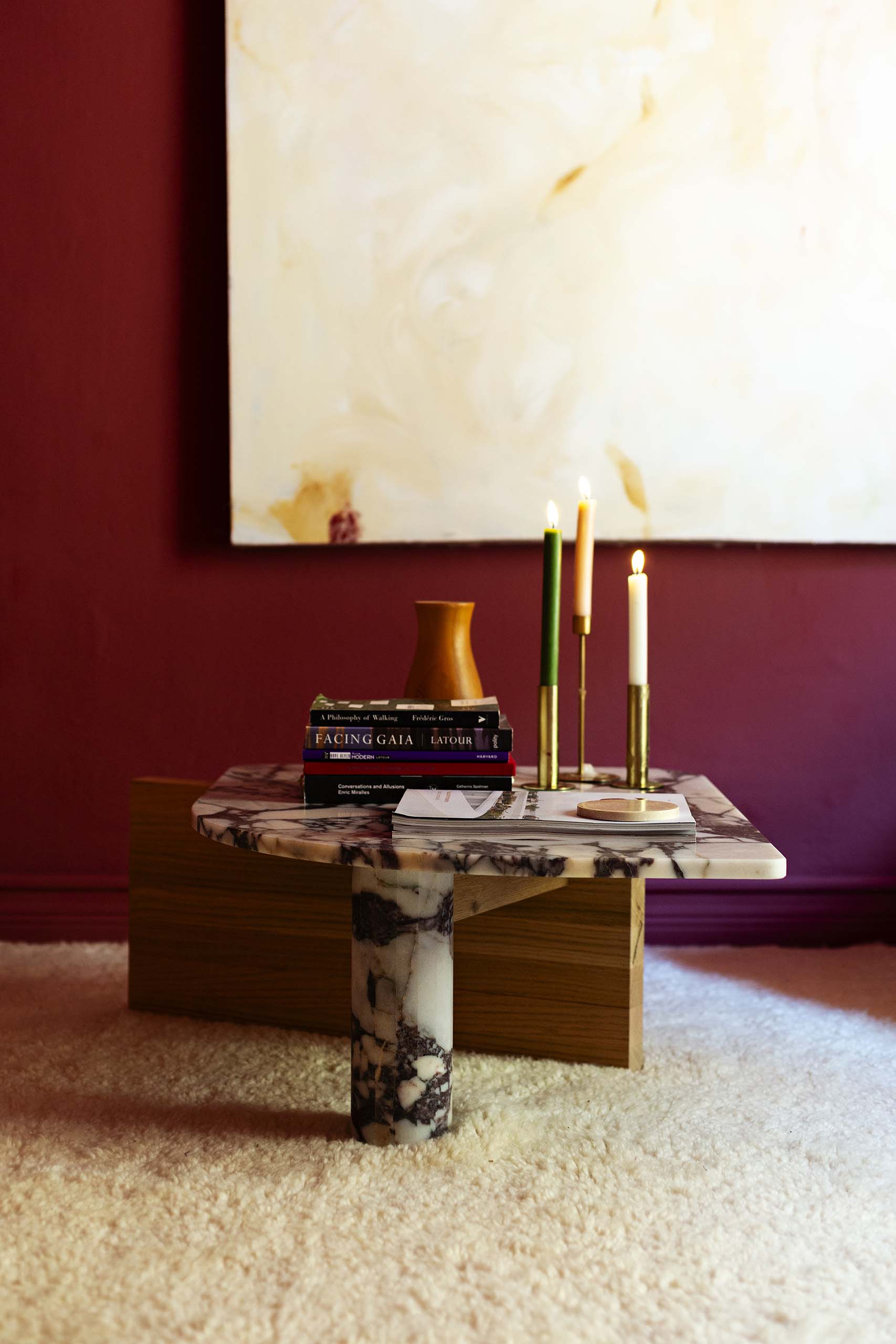Marble-top coffee table on beige carpet with three lit candles in brass holders, stacked books, and a wooden vase, against a burgundy wall with abstract painting.