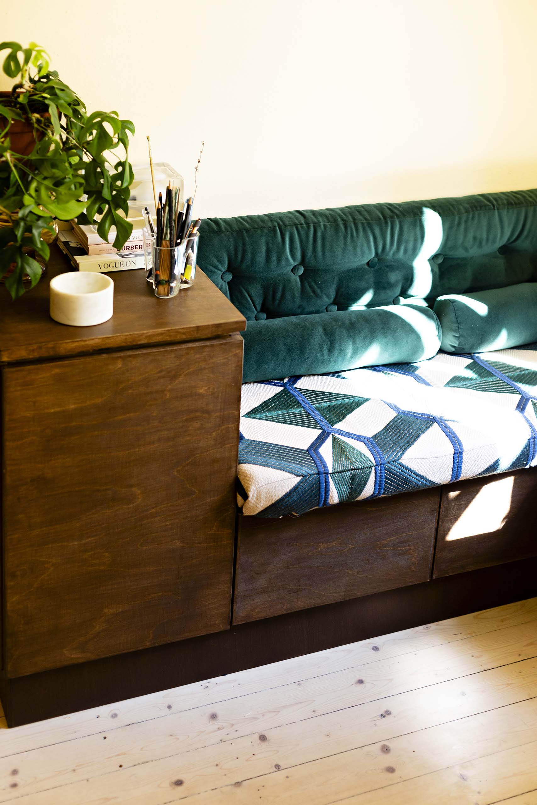 Cozy corner with a dark wooden bench featuring a green cushioned backrest and geometric cushion, next to a matching wooden side table holding a potted plant, books, and an assortment of pencils and paintbrushes.