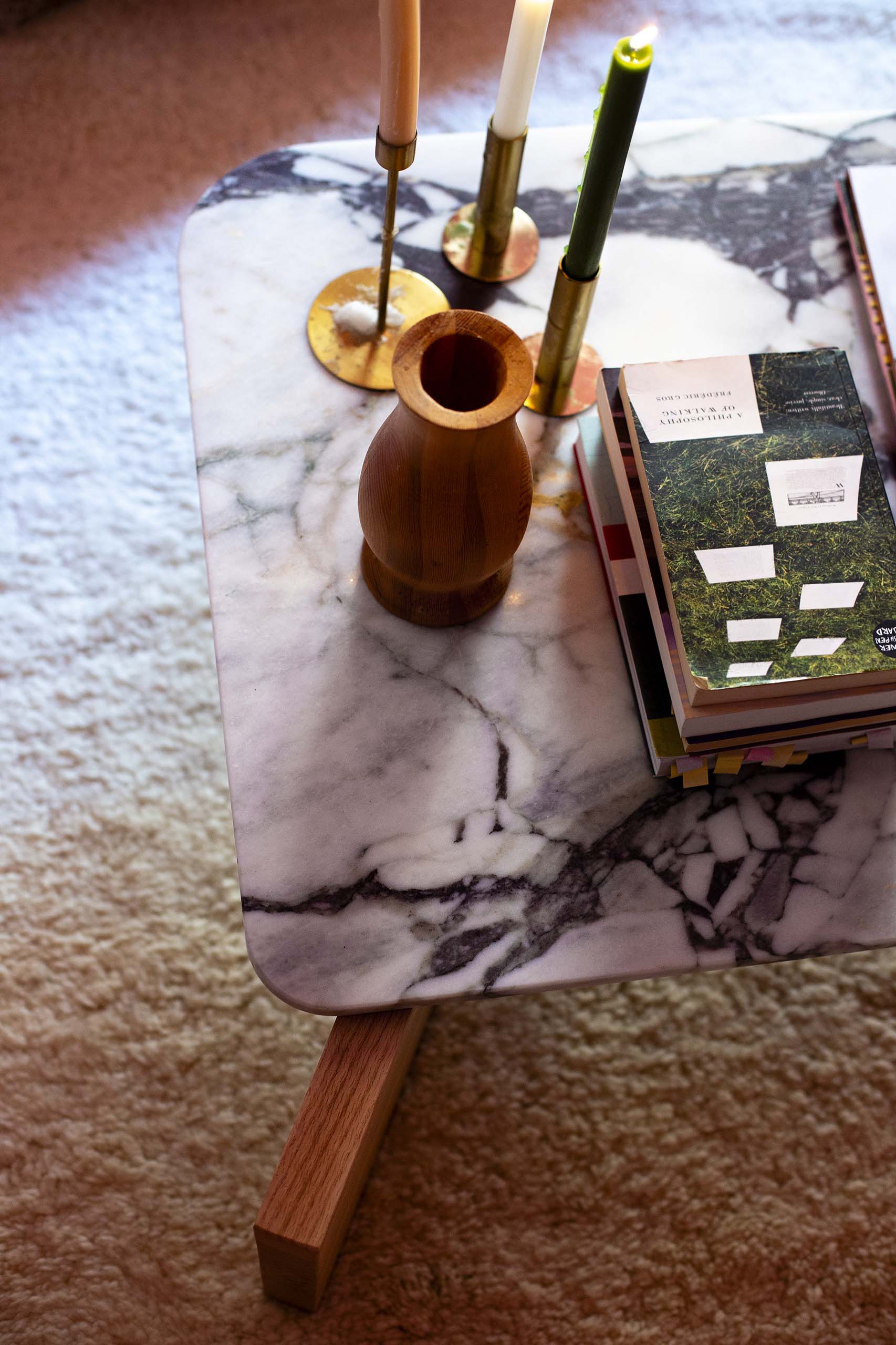 Marble-top coffee table with three brass candle holders holding different colored candles, a wooden vase, and a stack of books.