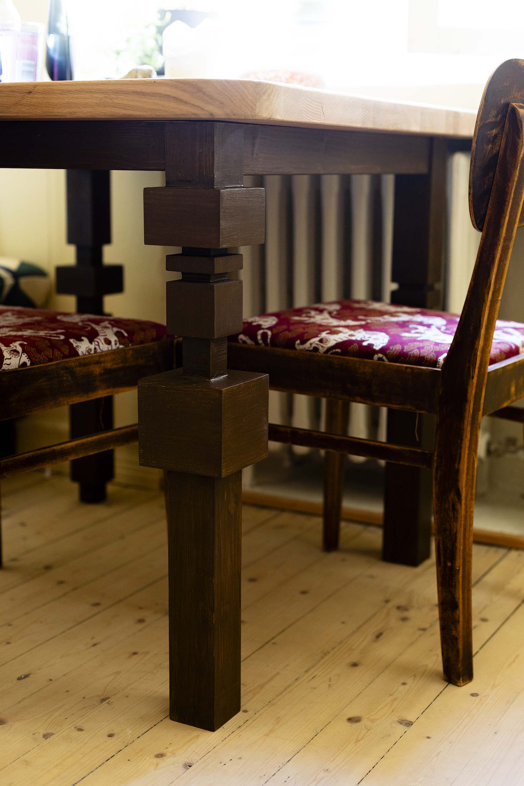 Close-up of a wooden table leg with a geometric design next to wooden chairs with red patterned cushions on a light wood floor.